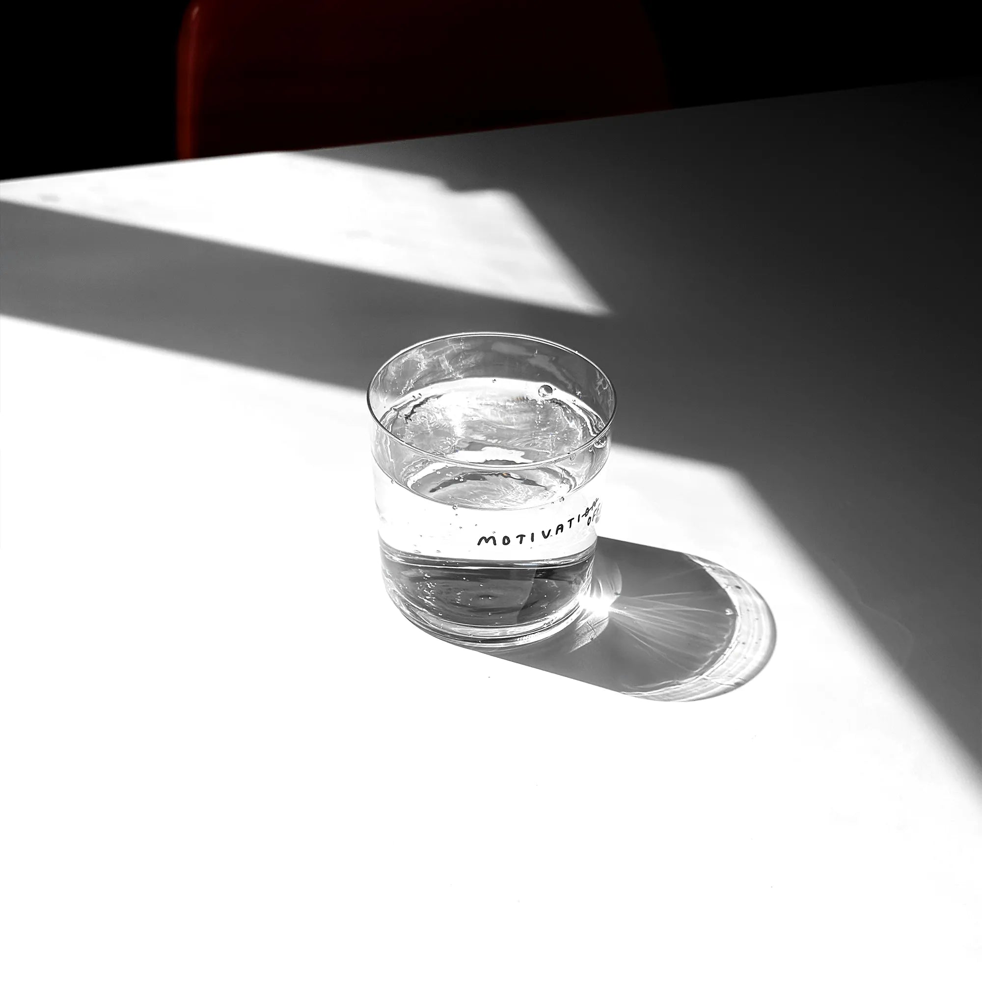 Großes Designer-Trinkglas mit sehr dünnen Glaswänden und einer schwarzen, einfarbigen Illustration auf der Vorderseite mit dem Motiv „MotivatioFF“. Das Wort Motivation steht ausgeschrieben aber ON ist durchgestrichen, darüber OFF. Illustration von Künstlerin, Illustratorin und Co-Gründerin Johanna Schwarzer.

Das Glas steht auf einem weißen Tisch, das Foto ist in einer Nahaufnahme, das Glas ist mit einer transparenten Flüssigkeit befüllt. Im Hintergrund viel Schatten. 