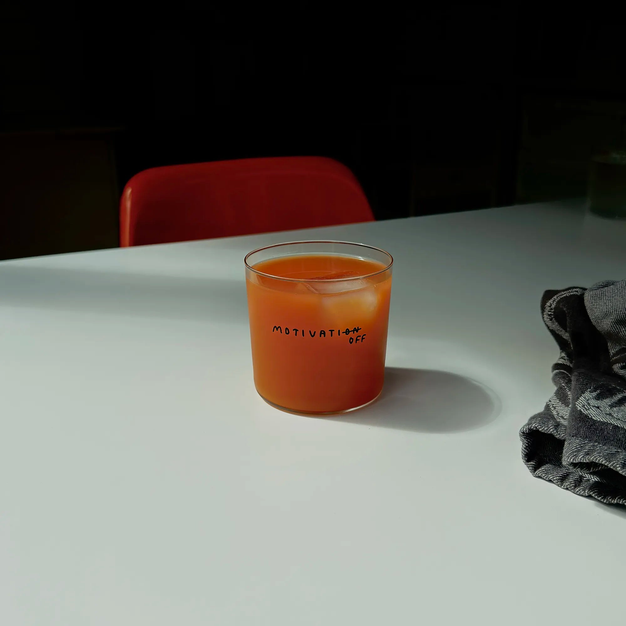Transparentes Designer Glass mit filigranen dünnen Wänden und Boden schwarzem Schriftzug "Motivatioff", wobei eigentlich Motivation geschrieben ist, das ON am Ende durchgestrichen ist, und durch OFF ersetzt wird. 

Das Glas ist mit einer orangenen Flüssigkeit und Eiswürfeln befüllt. Es steht auf einem weißen Tisch, ein graues Küchentuch liegt daneben, im Hintergrund steht ein orangener Stuhl.