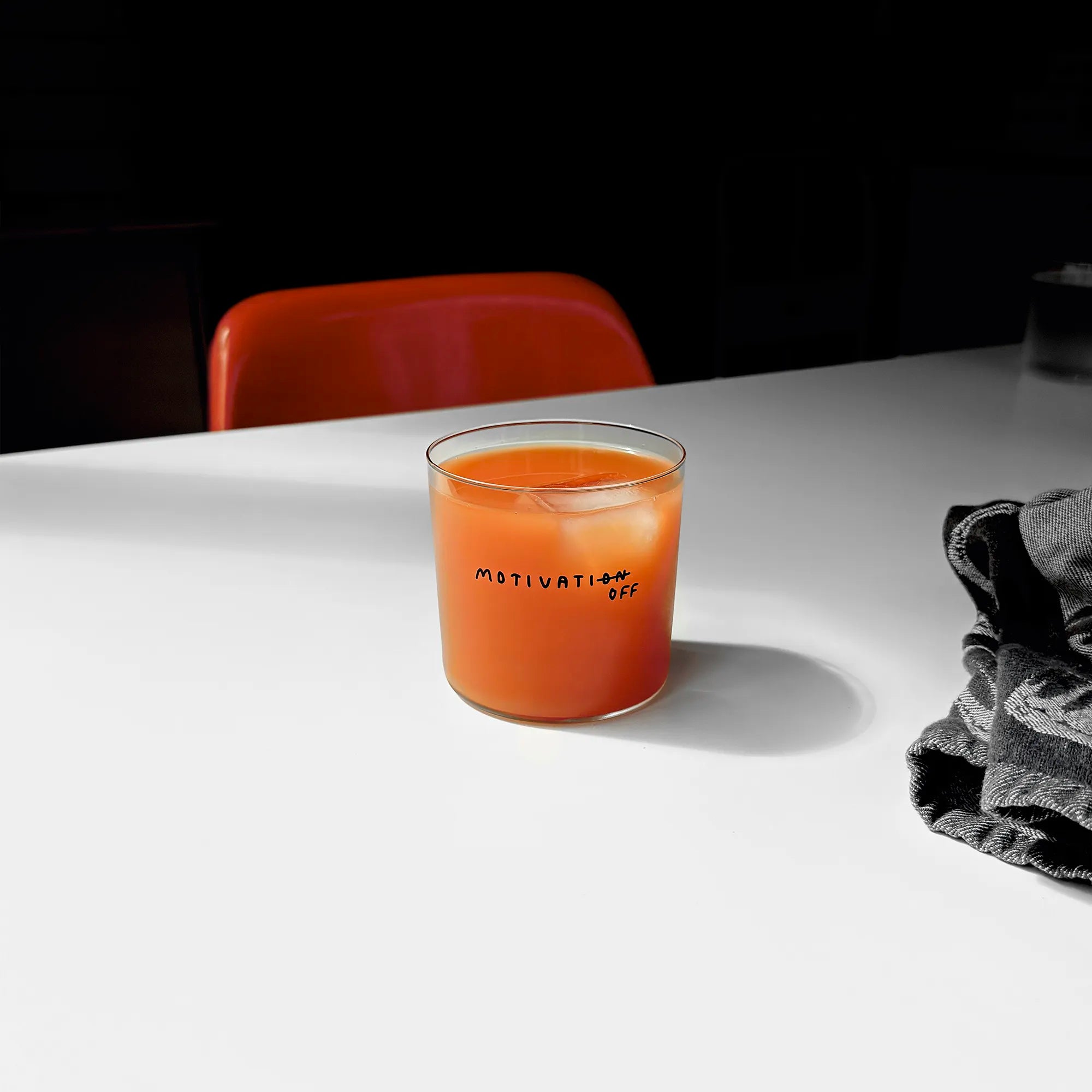 Transparentes Designer Glass mit filigranen dünnen Wänden und Boden schwarzem Schriftzug "Motivatioff", wobei eigentlich Motivation geschrieben ist, das ON am Ende durchgestrichen ist, und durch OFF ersetzt wird. 

Das Glas ist mit einer orangenen Flüssigkeit und Eiswürfeln befüllt. Es steht auf einem weißen Tisch, ein graues Küchentuch liegt daneben, im Hintergrund steht ein orangener Stuhl.