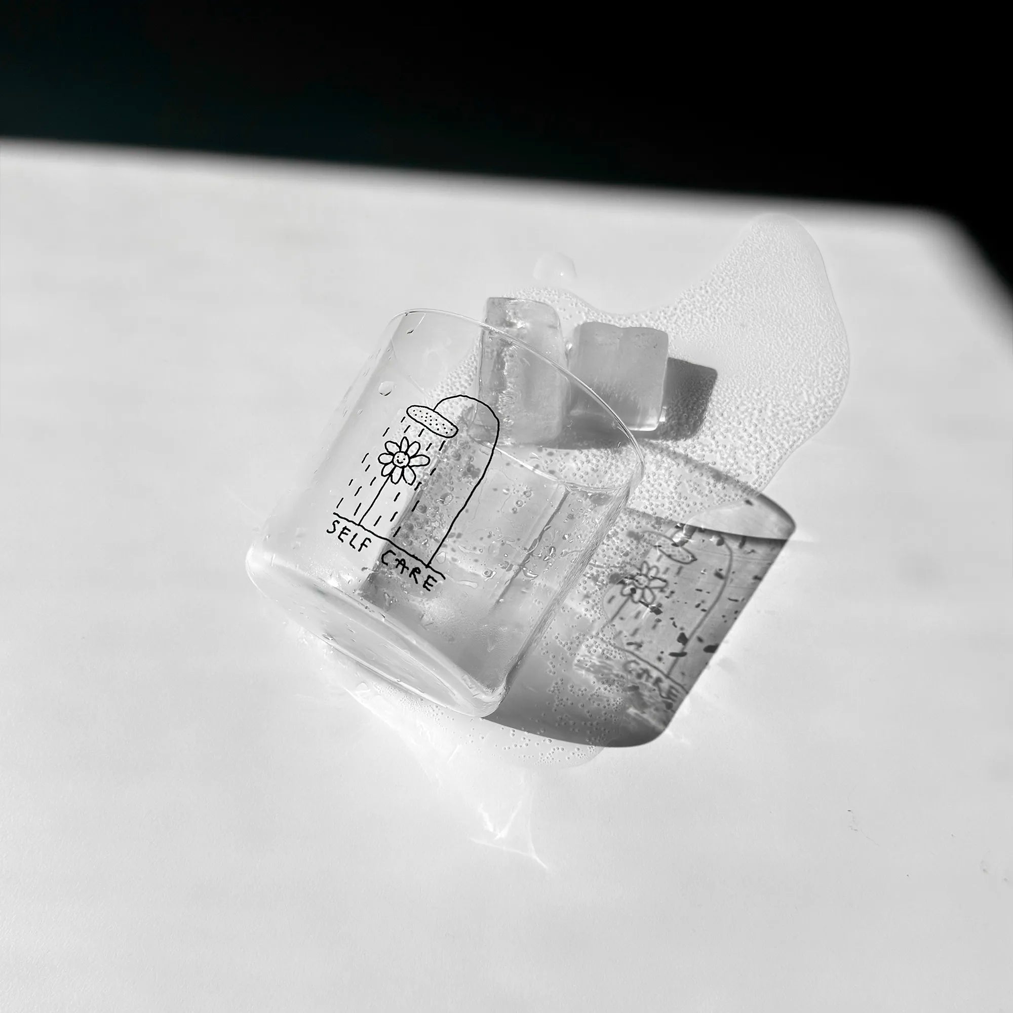 Transparentes Medium Designer Glas mit schwarzem Schriftzug "Self Care" und einer Illustration bei der eine Blume und einer Duschbrause steht und duscht, die Gläser haben dünnen Wänden und Boden. 
 
Das Glas ist mit einer transparenten Flüssigkeit befüllt und Eiswürfeln. Es liegt auf einem weißen Tisch wie umgeworfen, die Eiswürfel und Flüssigkeit sind ausgelaufen. Es wirft einen angenehmen Schatten. 