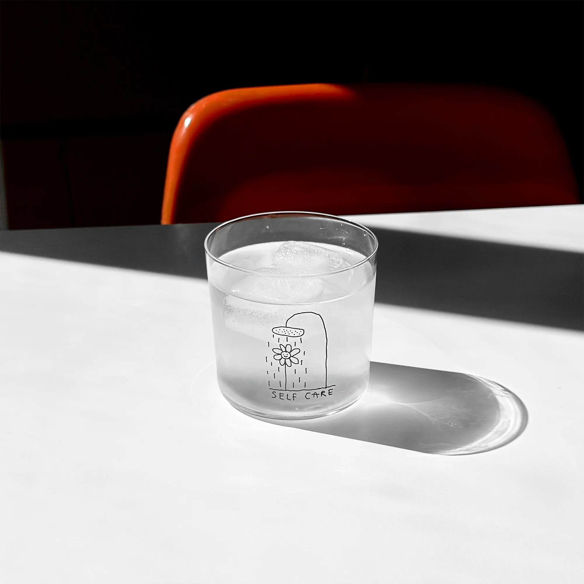 Transparentes Medium Designer Glas mit schwarzem Schriftzug "Self Care" und einer Illustration bei der eine Blume und einer Duschbrause steht und duscht, die Gläser haben dünnen Wänden und Boden. 
 
Das Glas ist mit einer transparentn Flüssigkeit und Eiswürfeln befüllt und steht auf einem weißen Tisch, daneben ein orangener Stuhl. 