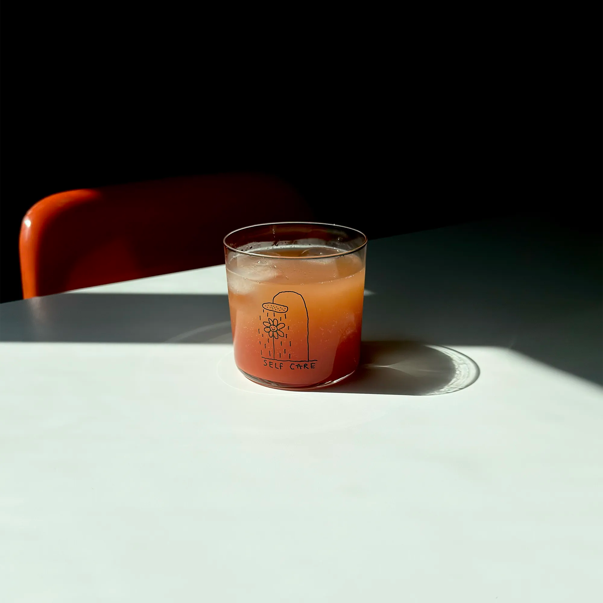 Transparentes Medium Designer Glas mit schwarzem Schriftzug "Self Care" und einer Illustration bei der eine Blume und einer Duschbrause steht und duscht, die Gläser haben dünnen Wänden und Boden. 
 
Das Glas ist mit einer orangenen Flüssigkeit und Eiswürfeln befüllt und steht auf einem weißen Tisch, daneben ein orangener Stuhl. 