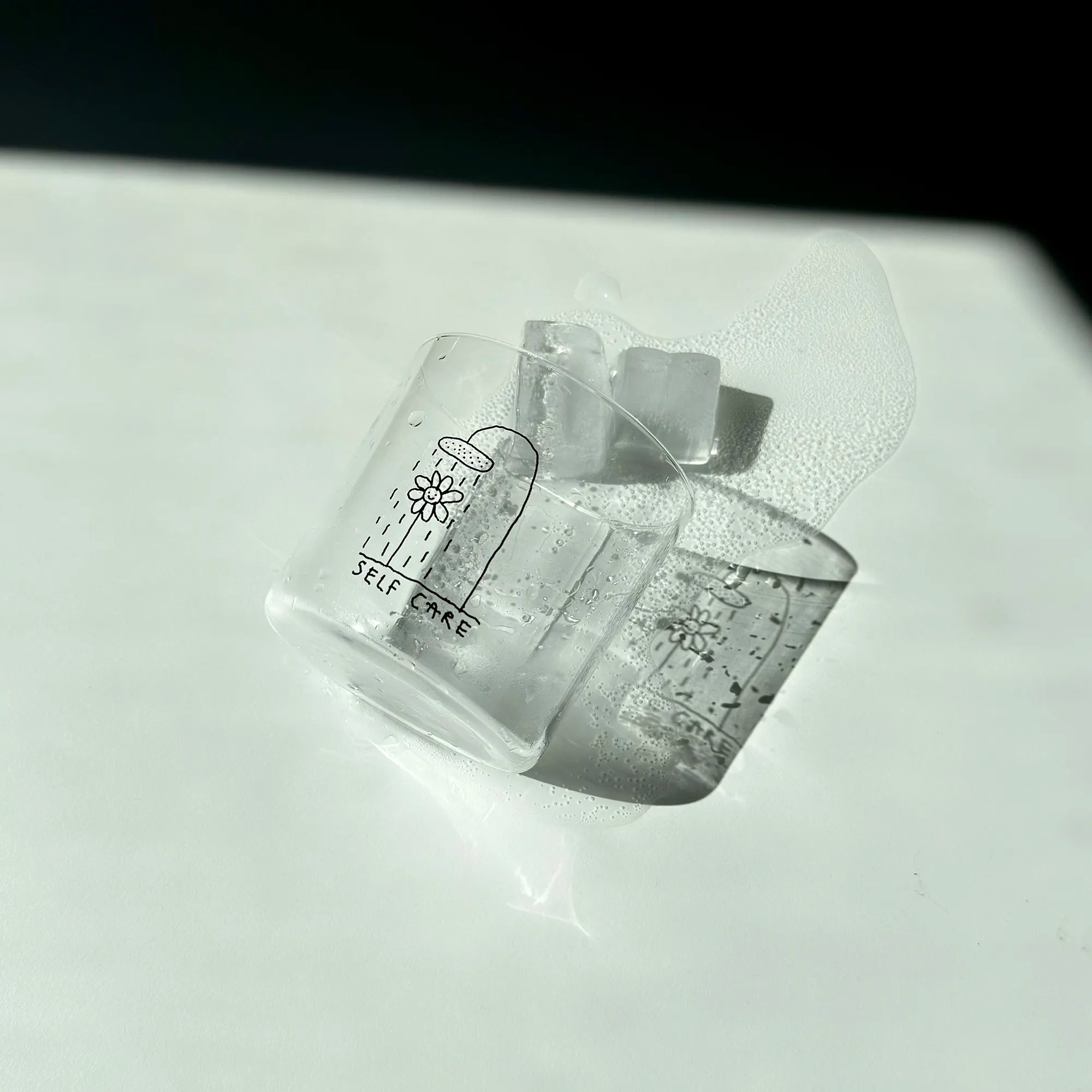 Transparentes Medium Designer Glas mit schwarzem Schriftzug "Self Care" und einer Illustration bei der eine Blume und einer Duschbrause steht und duscht, die Gläser haben dünnen Wänden und Boden. 
 
Das Glas ist mit einer transparenten Flüssigkeit befüllt und Eiswürfeln. Es liegt auf einem weißen Tisch wie umgeworfen, die Eiswürfel und Flüssigkeit sind ausgelaufen. Es wirft einen angenehmen Schatten. 