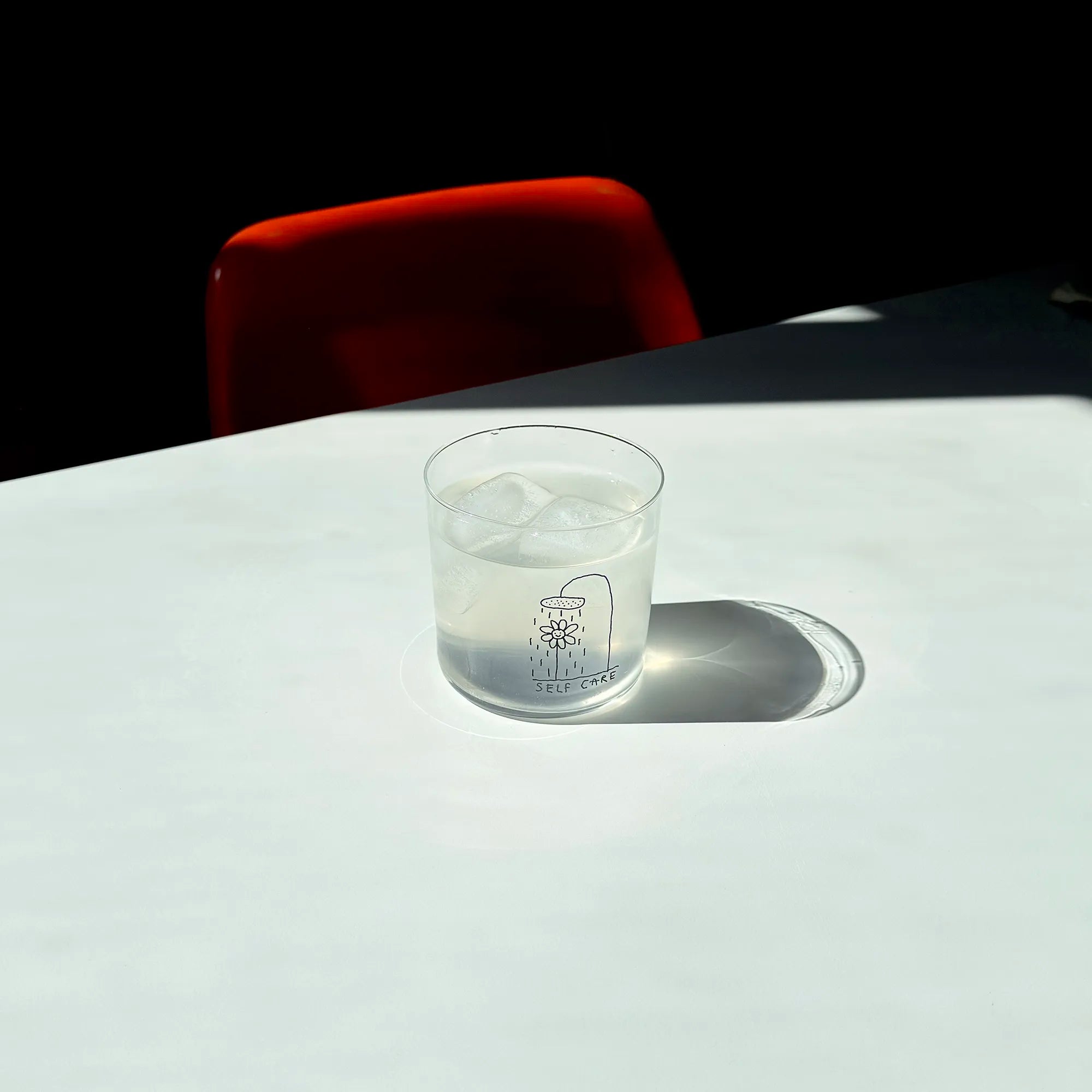 Transparentes Medium Designer Glas mit schwarzem Schriftzug "Self Care" und einer Illustration bei der eine Blume und einer Duschbrause steht und duscht, die Gläser haben dünnen Wänden und Boden. 
 
Das Glas ist mit einer transparentn Flüssigkeit und Eiswürfeln befüllt und steht auf einem weißen Tisch, daneben ein orangener Stuhl. 