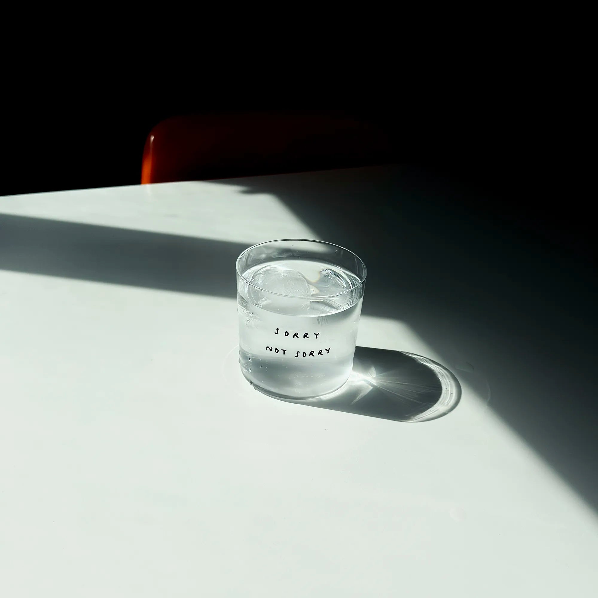 Medium Designer-Trinkglas mit sehr dünnen Glaswänden und einer schwarzen, einfarbigen Illustration auf der Vorderseite. Das Motiv zeigt eine Typografie in der geschrieben ist SORRY NOT SORRY.

Das Glas steht auf einem weißen Tisch in angenehmen Licht, dahinter steht ein orangener Stuhl. Das Glas ist gefüllt mit einer transparenten Flüssigkeit und einem Eiswürfel