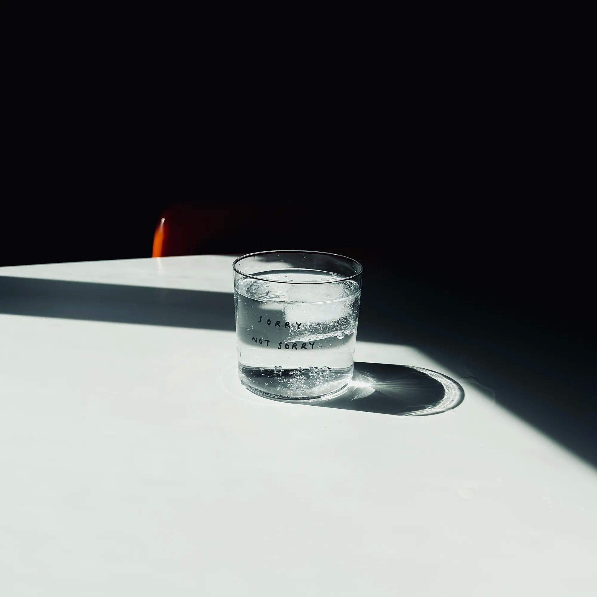 Medium Designer-Trinkglas mit sehr dünnen Glaswänden und einer schwarzen, einfarbigen Illustration auf der Vorderseite. Das Motiv zeigt eine Typografie in der geschrieben ist SORRY NOT SORRY.

Das Glas steht auf einem weißen Tisch in angenehmen Licht, dahinter steht ein orangener Stuhl. Das Glas ist gefüllt mit einer transparenten Flüssigkeit und einem Eiswürfel