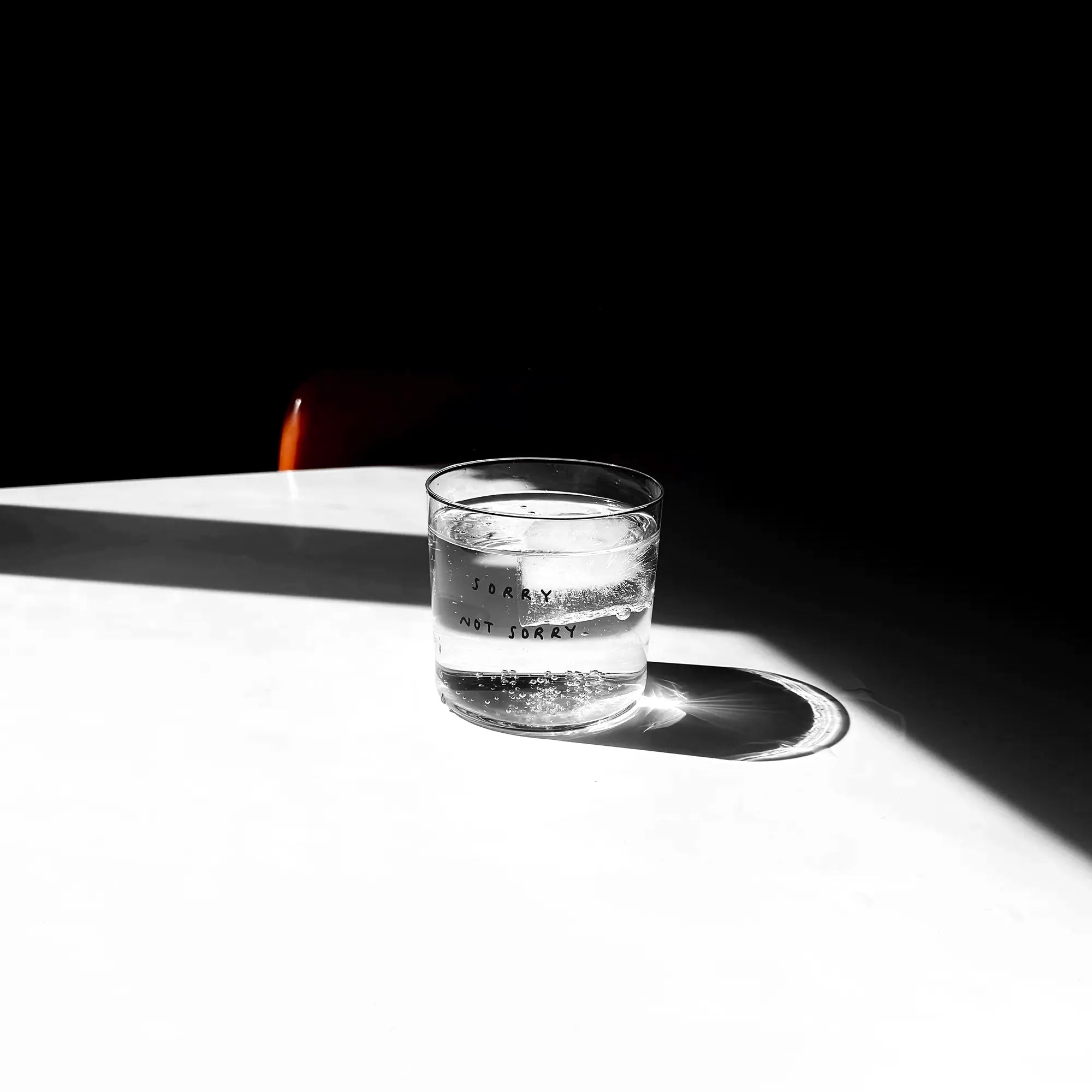 Medium Designer-Trinkglas mit sehr dünnen Glaswänden und einer schwarzen, einfarbigen Illustration auf der Vorderseite. Das Motiv zeigt eine Typografie in der geschrieben ist SORRY NOT SORRY.

Das Glas steht auf einem weißen Tisch in angenehmen Licht, dahinter steht ein orangener Stuhl. Das Glas ist gefüllt mit einer transparenten Flüssigkeit und einem Eiswürfel