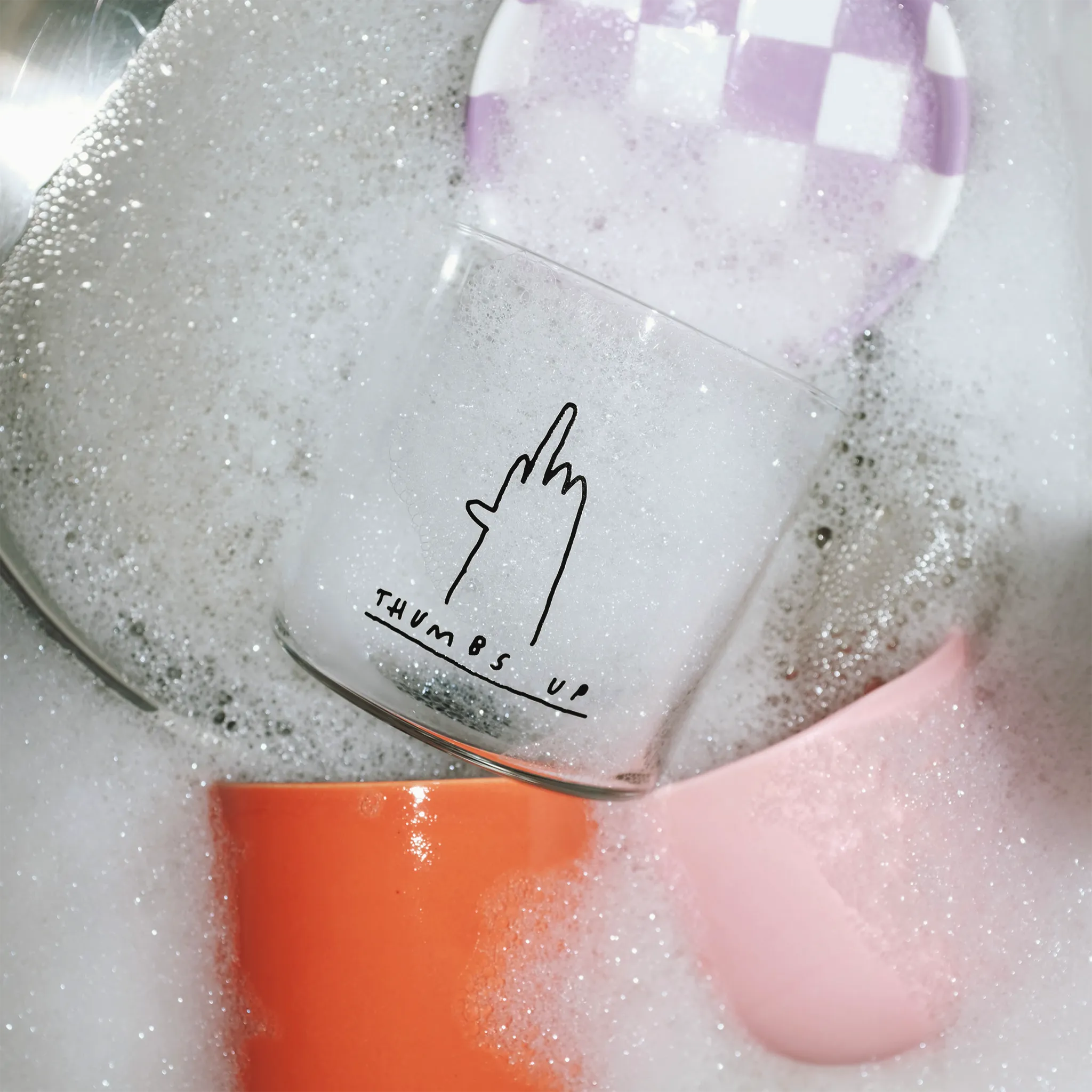 Medium Designer-Trinkglas mit sehr dünnen Glaswänden und einer schwarzen, einfarbigen Illustration auf der Vorderseite. Das Motiv zeigt eine Typografie in der geschrieben THUMBS UP und einer Illustration einer Hand die den Mittelfinger zeigt.

Das Glas ist in einer Spüle mit viel Schaum um sich herum, wird mit Wasser befüllt.