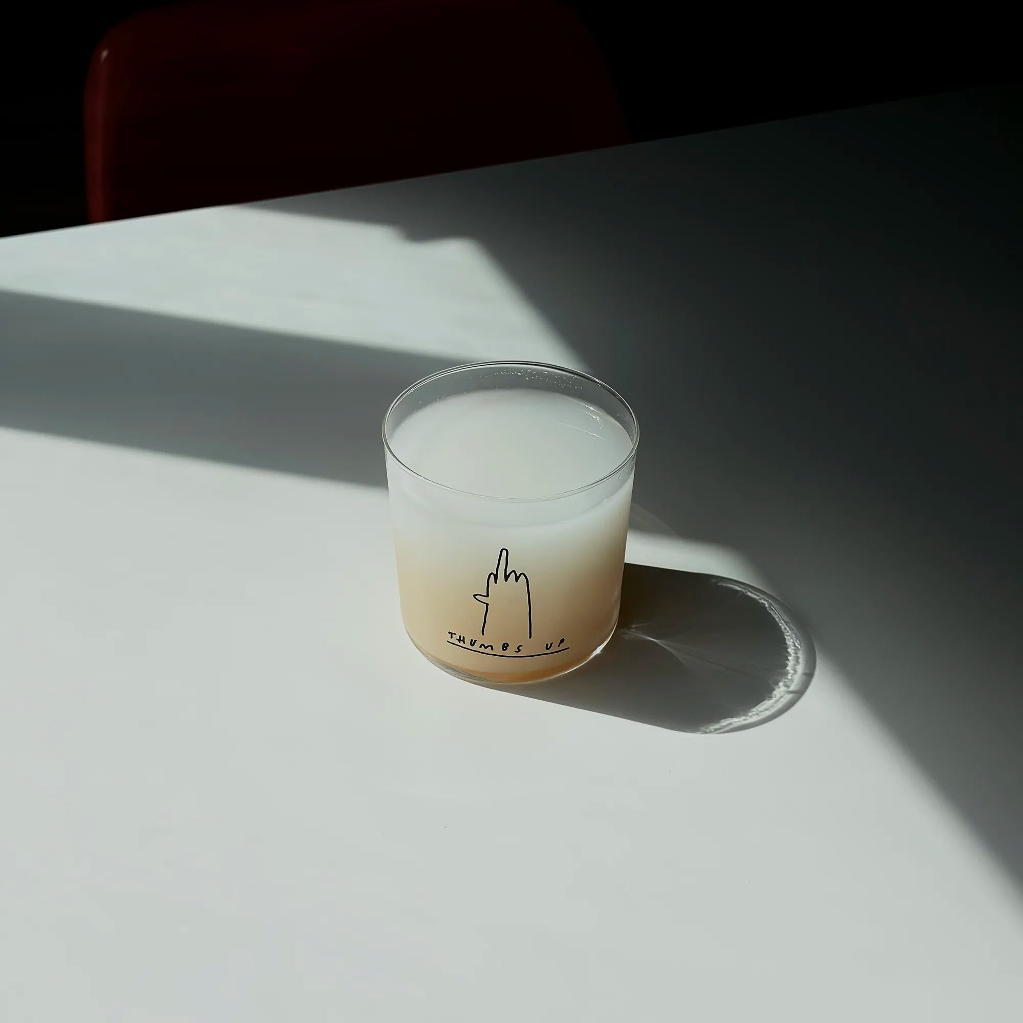 Medium Designer-Trinkglas mit sehr dünnen Glaswänden und einer schwarzen, einfarbigen Illustration auf der Vorderseite. Das Motiv zeigt eine Typografie in der geschrieben THUMBS UP und einer Illustration einer Hand die den Mittelfinger zeigt.

Das Glas ist mit einer weißen Flüssigkeit befüllt, steht auf einem Weißen Tisch in einem Lichtkegel, dahinter ist ein Stuhl ganz dezent erkennbar. 