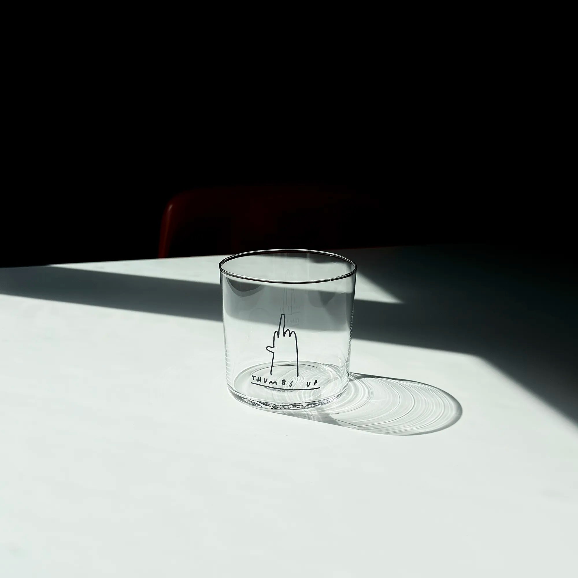 Medium Designer-Trinkglas mit sehr dünnen Glaswänden und einer schwarzen, einfarbigen Illustration auf der Vorderseite. Das Motiv zeigt eine Typografie in der geschrieben THUMBS UP und einer Illustration einer Hand die den Mittelfinger zeigt.

Das Glas ist ungefüllt, steht auf einem Weißen Tisch in einem Lichtkegel, dahinter ist ein Stuhl ganz dezent erkennbar. 