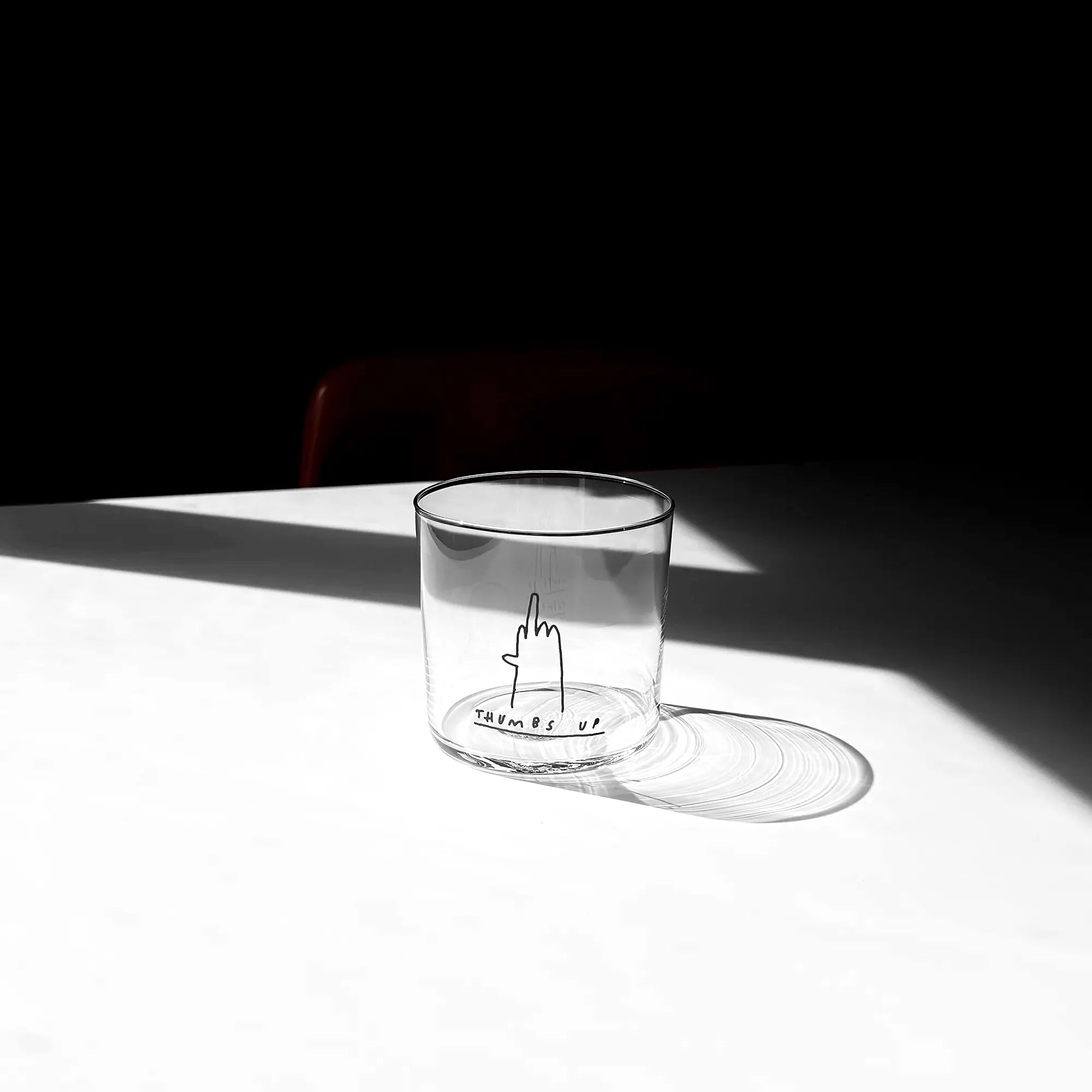 Medium Designer-Trinkglas mit sehr dünnen Glaswänden und einer schwarzen, einfarbigen Illustration auf der Vorderseite. Das Motiv zeigt eine Typografie in der geschrieben THUMBS UP und einer Illustration einer Hand die den Mittelfinger zeigt.

Das Glas ist ungefüllt, steht auf einem Weißen Tisch in einem Lichtkegel, dahinter ist ein Stuhl ganz dezent erkennbar. 