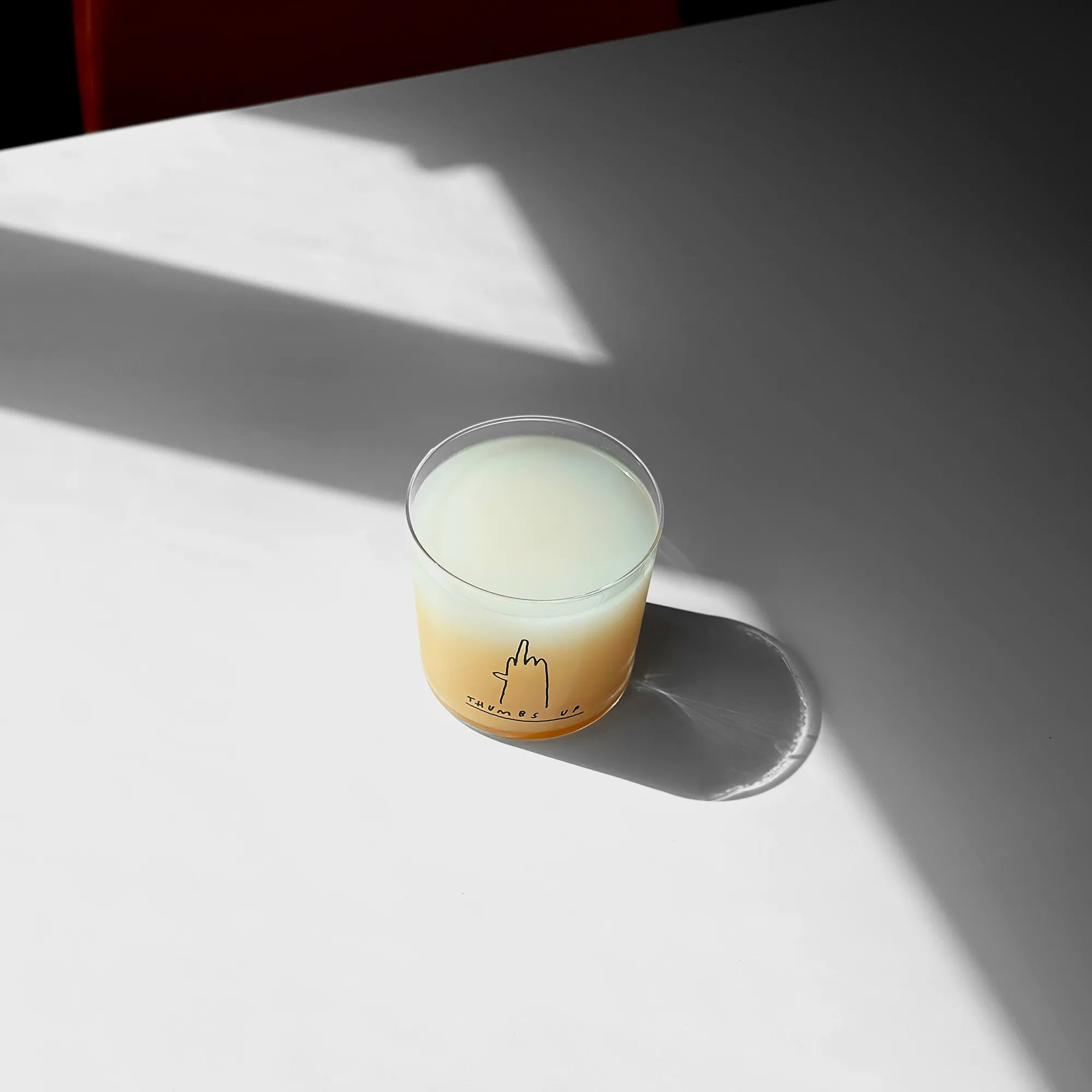 Medium Designer-Trinkglas mit sehr dünnen Glaswänden und einer schwarzen, einfarbigen Illustration auf der Vorderseite. Das Motiv zeigt eine Typografie in der geschrieben THUMBS UP und einer Illustration einer Hand die den Mittelfinger zeigt.

Das Glas ist mit einer weißen Flüssigkeit befüllt, steht auf einem Weißen Tisch in einem Lichtkegel, dahinter ist ein Stuhl ganz dezent erkennbar. 