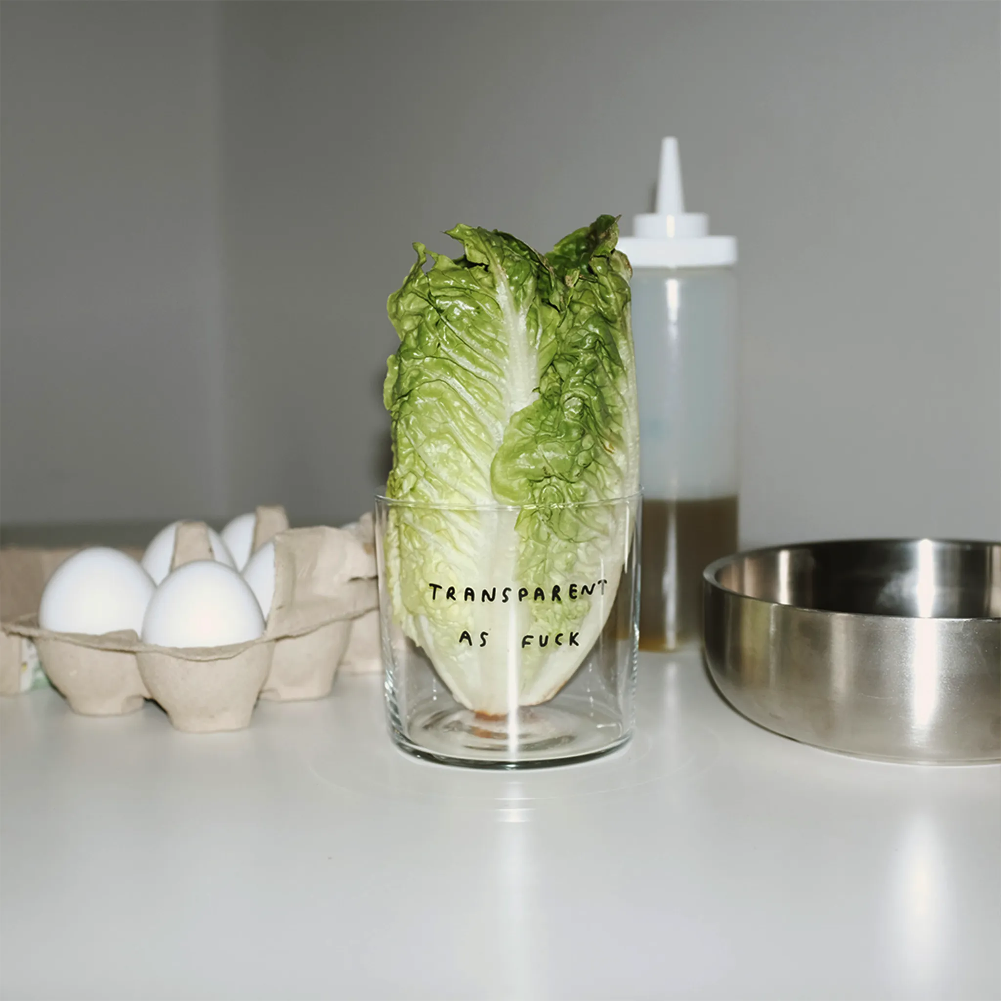 Medium Designer-Trinkglas mit sehr dünnen Glaswänden und einer schwarzen, einfarbigen Illustration auf der Vorderseite. Das Motiv zeigt eine Typografie in der geschrieben ist TRANSPARENT AS FUCK.

Das Glas steht auf einem weißen Tisch, im Hintergrund sieht man Eier, eine Metallschale und eine Flasche. In dem Glas steckt ein Salatherz.