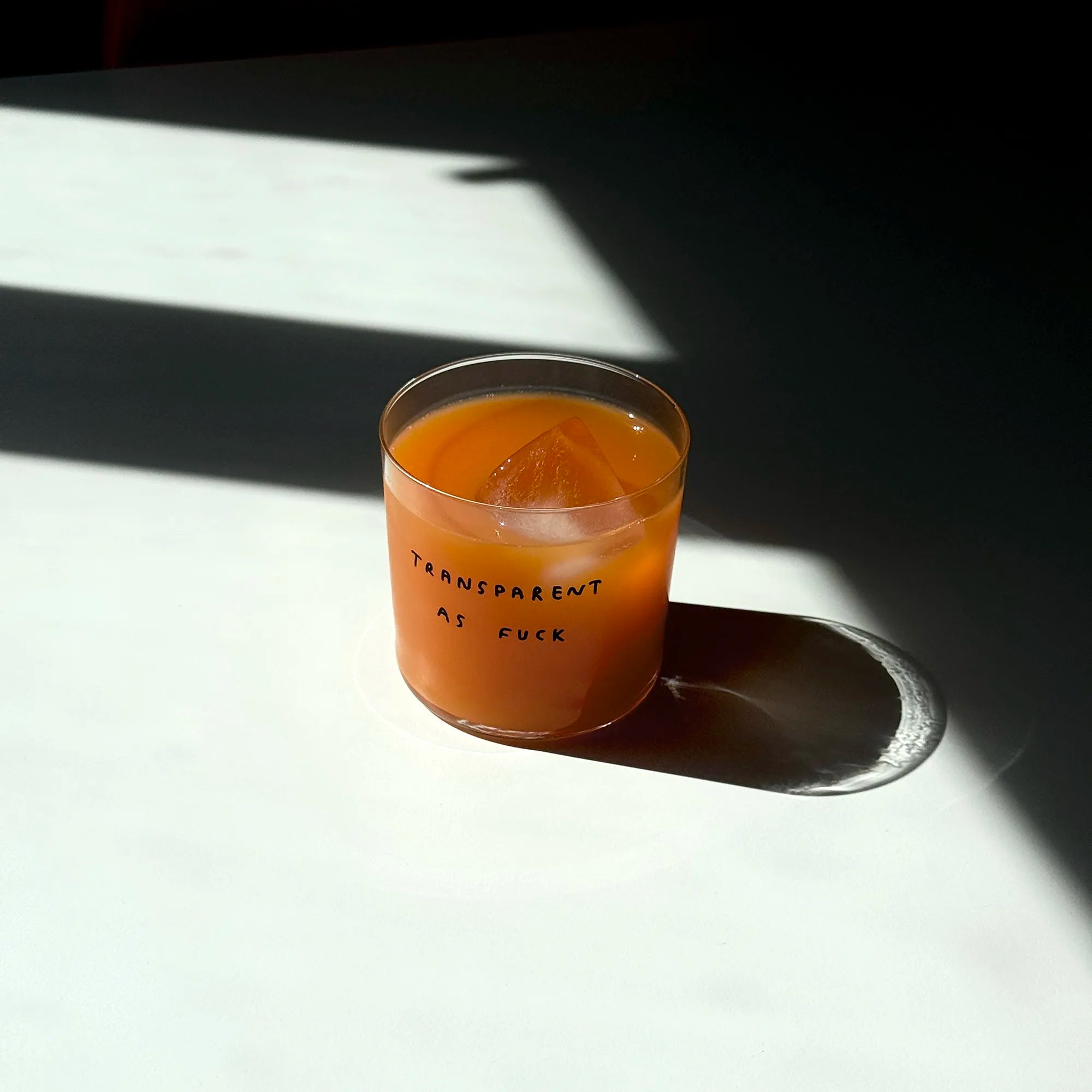 Medium Designer-Trinkglas mit sehr dünnen Glaswänden und einer schwarzen, einfarbigen Illustration auf der Vorderseite. Das Motiv zeigt eine Typografie in der geschrieben ist TRANSPARENT AS FUCK.

Das Glas steht auf einem weißen Tisch und ist mit einer Orangenen Flüssigkeit gefüllt. Ein Eiswürfel ist im Glas, das Glas wirft einen großen Schatten. 