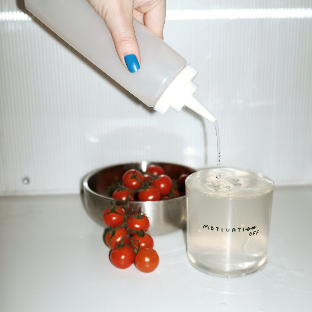 Transparentes Designer Glass mit filigranen dünnen Wänden und Boden schwarzem Schriftzug "Motivatioff", wobei eigentlich Motivation geschrieben ist, das ON am Ende durchgestrichen ist, und durch OFF ersetzt wird. Weißer Hintergrund.

Das Glas steht auf einem weißen Tisch, wird mit einer Flüssigkeit befüllt. Eine Aluminium Schale mit Tomaten steht im Hintergrund.