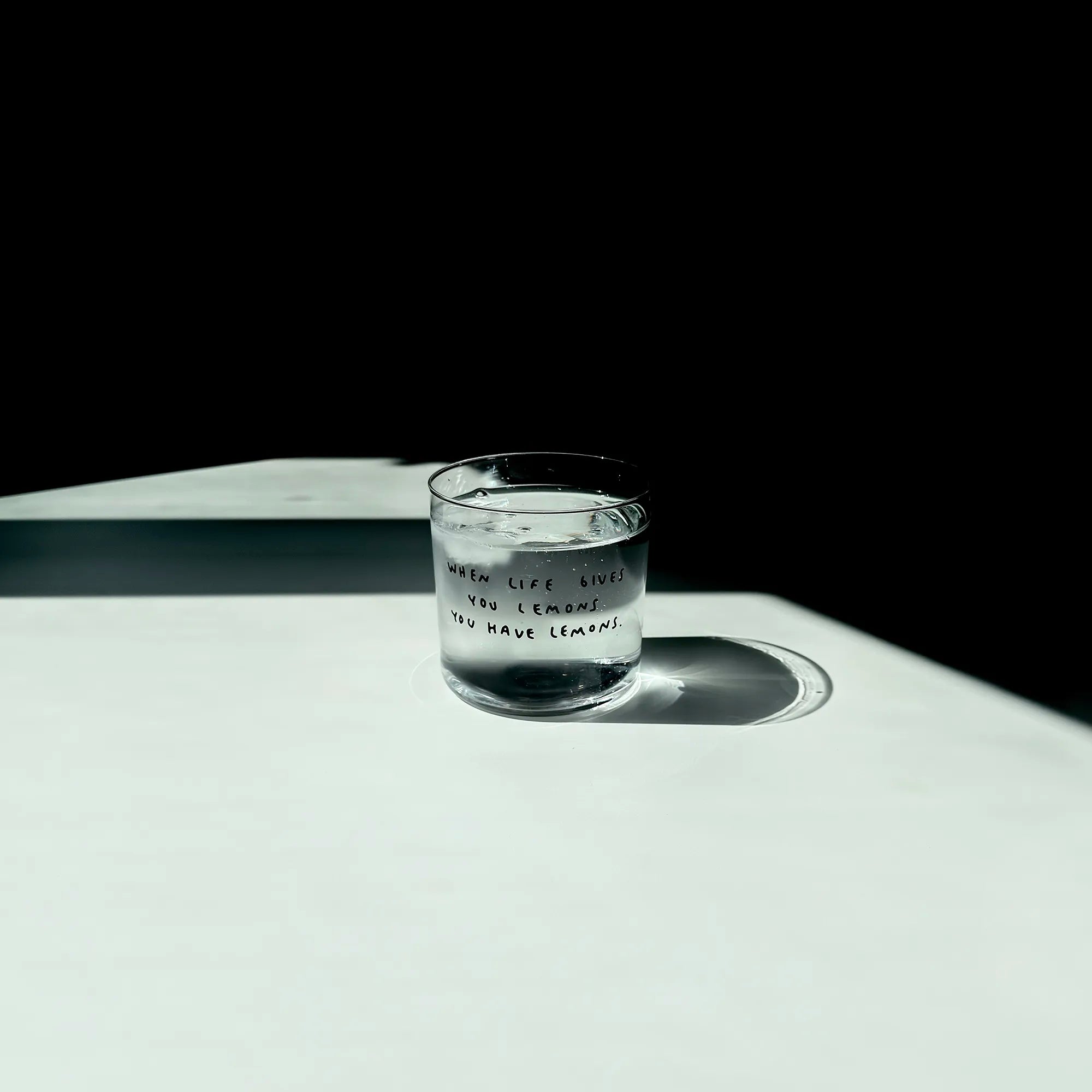 Transparentes Medium Designer Glas mit schwarzem Schriftzug "when life gives you lemons" mit dünnen Wänden und Boden. Das Glas steht im Licht auf einem weißen Tisch und wirft einen weiten Schatten. Das Glas ist mit einer transparenten Flüssigkeit und Eiswürfeln befüllt. 