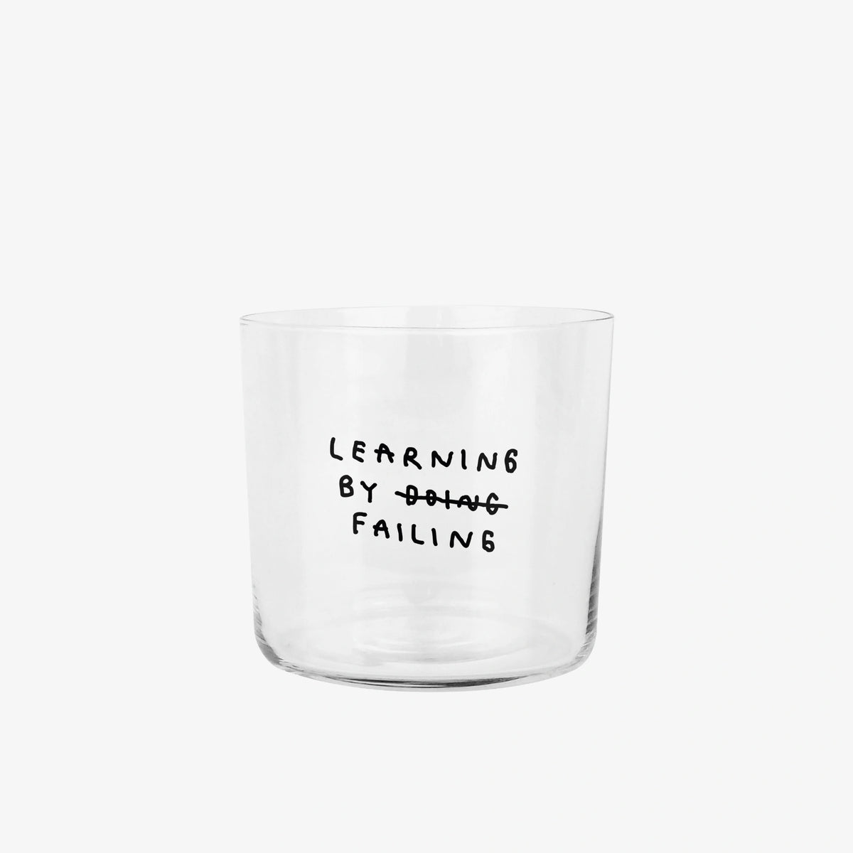 Klares, zylindrisches Trinkglas mit schwarzem, handschriftlich wirkendem Text. Darauf steht „Learning by doing“, wobei das Wort „doing“ durchgestrichen ist und darunter „failing“ steht, sodass der Satz humorvoll zu „Learning by failing“ geändert wird.