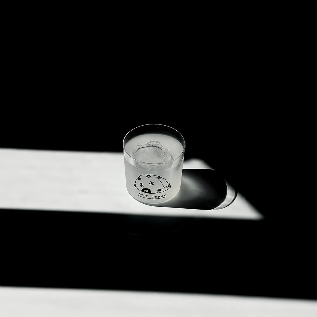 Transparentes Medium Designer Glas mit schwarzem Schriftzug "Not Today". Über dem Schriftzug ist eine Illustration bei der zwei Augen unter einer Art Decke hervorschauen. Das Glas hat dünnen Wänden und Boden. Das Glas steht im Licht auf einem weißen Tisch. Der Rest des Bildes ist dunkel in Schatten gefallen.

Das Glas ist befüllt mit einer klaren Flüssigkeit und einem Eiswürfel.