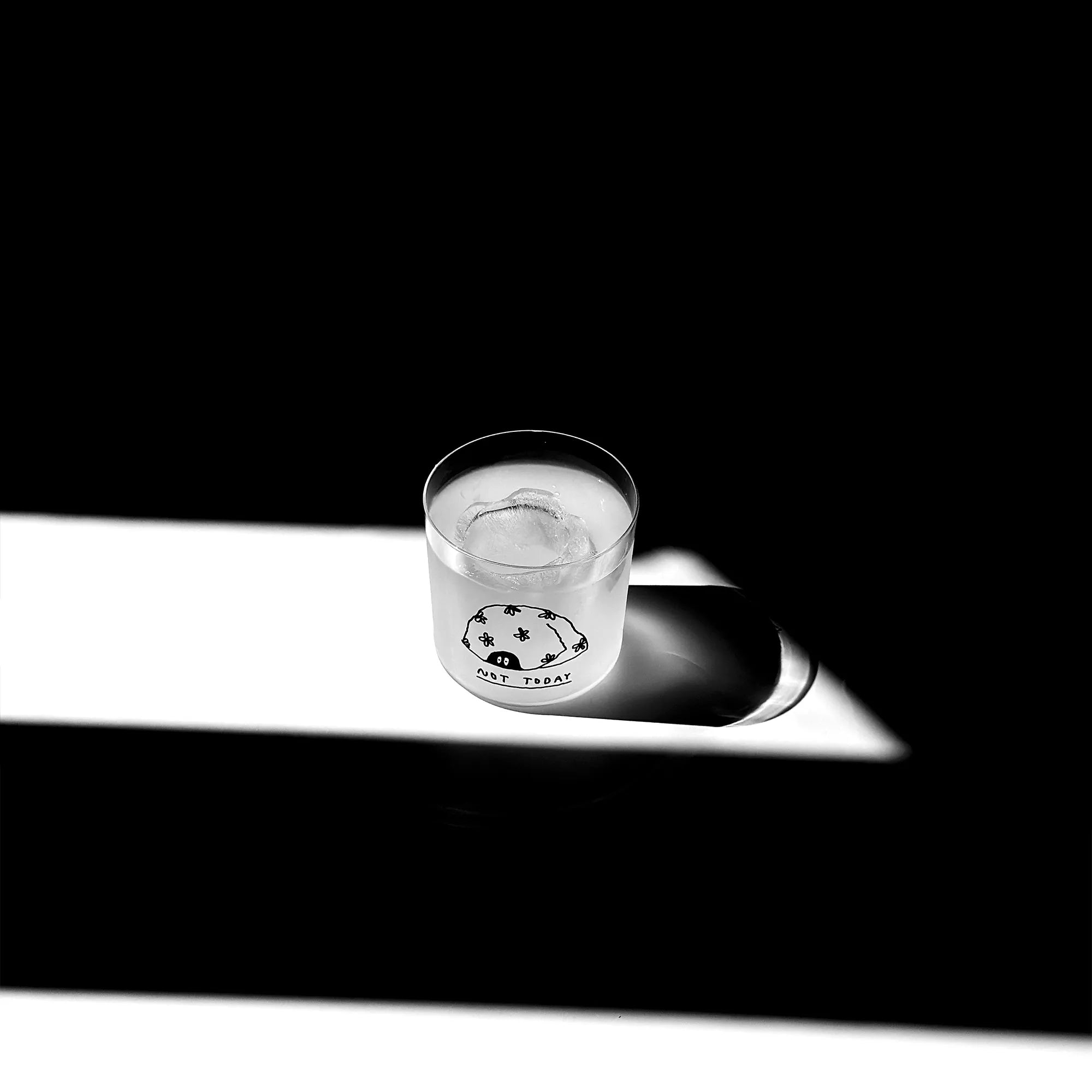 Transparentes Medium Designer Glas mit schwarzem Schriftzug "Not Today". Über dem Schriftzug ist eine Illustration bei der zwei Augen unter einer Art Decke hervorschauen. Das Glas hat dünnen Wänden und Boden. Das Glas steht im Licht auf einem weißen Tisch. Der Rest des Bildes ist dunkel in Schatten gefallen.

Das Glas ist befüllt mit einer klaren Flüssigkeit und einem Eiswürfel.