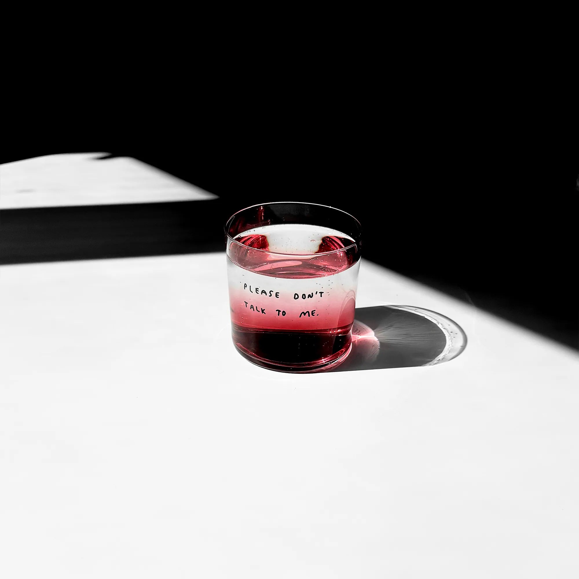 Medium Designer-Trinkglas mit sehr dünnen Glaswänden und einer schwarzen, einfarbigen Illustration auf der Vorderseite. Das Motiv zeigt eine Typografie in der geschrieben ist PLEASE DON'T TALK TO ME.

Das Glas ist mit einer roten leicht transparenten Flüssigkeit gefüllt und steht auf einem weißen Tisch. 