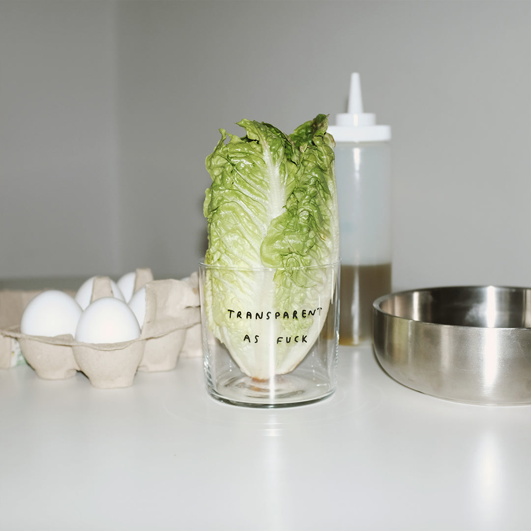 Medium Designer-Trinkglas mit sehr dünnen Glaswänden und einer schwarzen, einfarbigen Illustration auf der Vorderseite. Das Motiv zeigt eine Typografie in der geschrieben ist TRANSPARENT AS FUCK.

Das Glas steht auf einem weißen Tisch, im Hintergrund sieht man Eier, eine Metallschale und eine Flasche. In dem Glas steckt ein Salatherz.