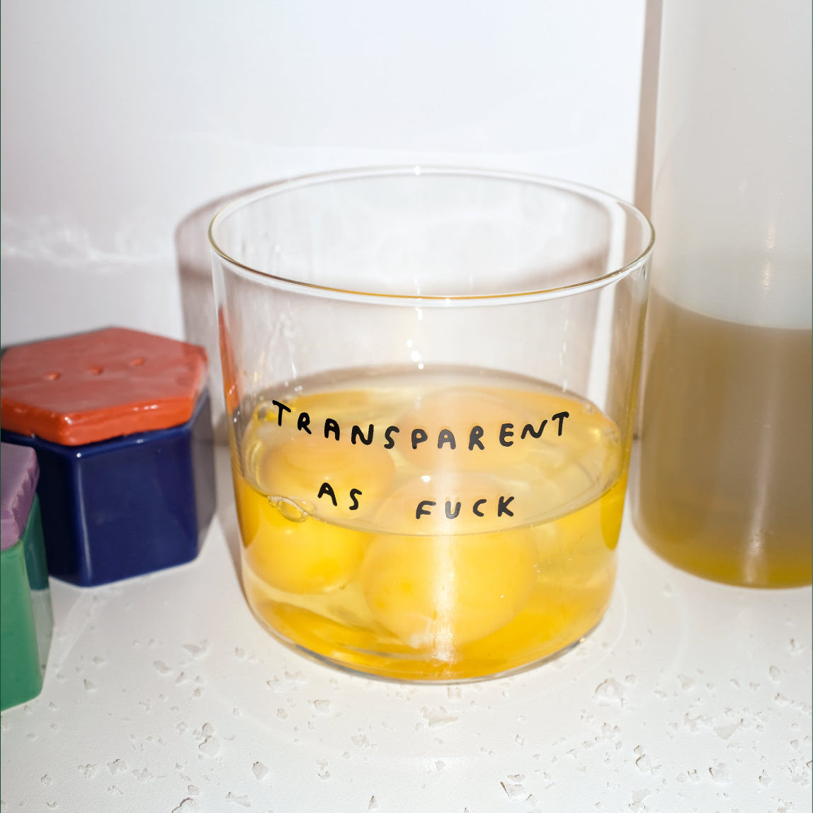 Transparentes Designer Glas mit dem Motiv transparent as fuck. Das Glas ist gefüllt mit einer gelben Flüssigkeit und steht auf einer Küchenfront.