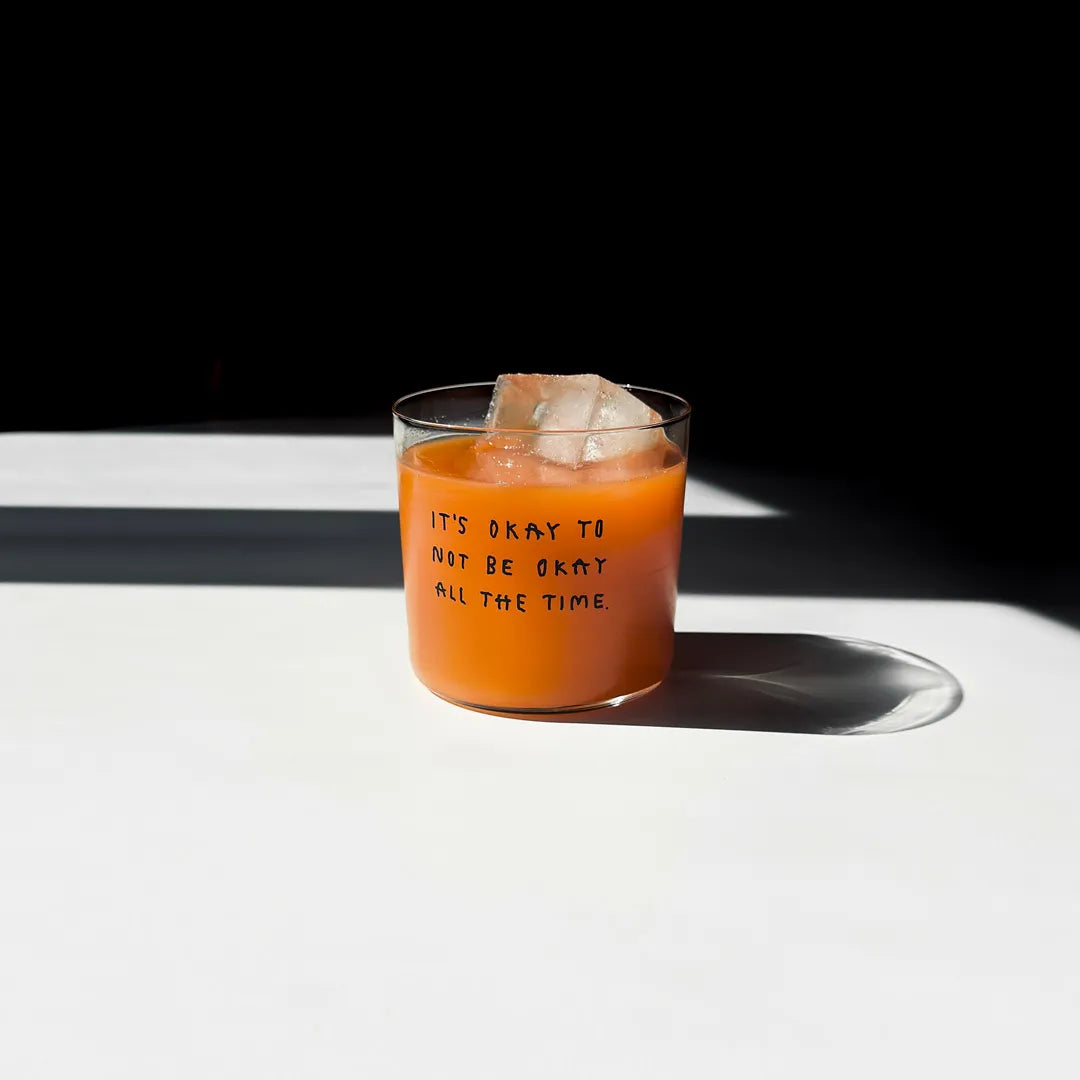 Transparentes Medium Designer Glas mit schwarzem Schriftzug "It's okay to not be okay all the time" mit dünnen Wänden und Boden. 

Das Glas ist mit einer orangenen Flüssigkeit befüllt und Eiswürfeln. Es steht auf einem weißen Tisch und wirft einen schatten.