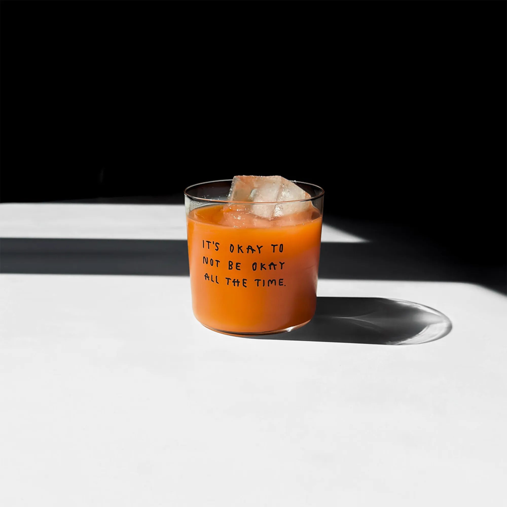 Transparentes Medium Designer Glas mit schwarzem Schriftzug "It's okay to not be okay all the time" mit dünnen Wänden und Boden. 

Das Glas ist mit einer orangenen Flüssigkeit befüllt und Eiswürfeln. Es steht auf einem weißen Tisch und wirft einen schatten.
