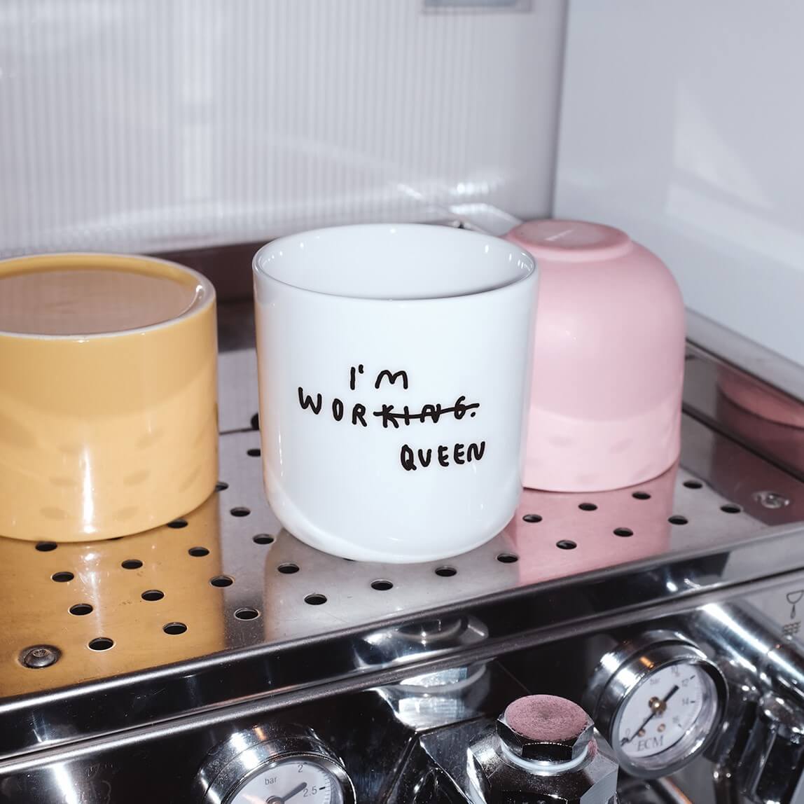 Weißer henkelloser Porzellan-Becher mit einer minimalistischen schwarzen Illustration  in der in Handschriftartiger Typografie I'M WORKING steht. Das KING wurde durchgestrichen und unten drunter wurde QUEEN gestrichen. 

Der Becher steht auf einer Kaffeemaschine neben zwei anderen Tassen.
