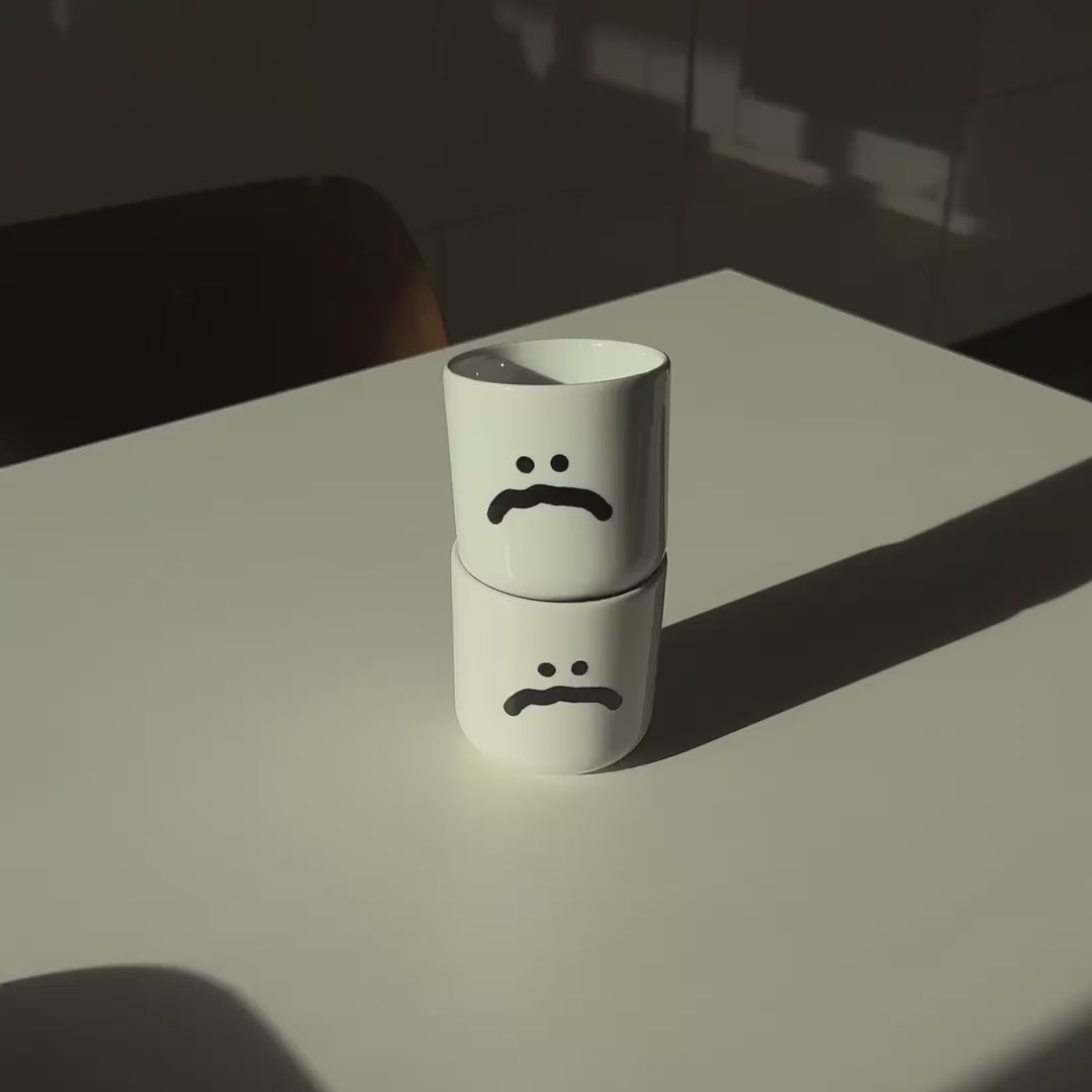 Zwei weiße henkelloser Porzellan-Becher mit einer einfachen schwarzen Illustration eines großen grinsenden Smiley und einem großen traurigen Smiley.

Beide Becher stehen gestapelt auf einem weißen Tisch mit Holzstuhl im Hintergrund. In dem Video wird die obere Tasse gedreht so dass das traurige Smiley nicht mehr sichtbar ist und das lachende Smiley von der anderen Seite gezeigt wird. 