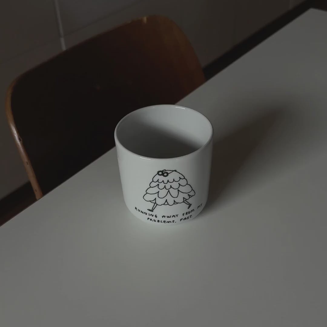 Weißer henkelloser Porzellan-Becher mit einer einfachen schwarzen Illustration einer kleinen, flauschigen, cartoonartigen Figur mit Beinen, die davonläuft. Unter der Zeichnung steht in handschriftlicher Schrift der Satz: „Running away from my problems, fast.“ (Deutsch: „Ich renne schnell vor meinen Problemen davon.“).

Die Tasse wird auf einem weißen Tisch stehend von einer Hand positioniert und leicht gedreht.