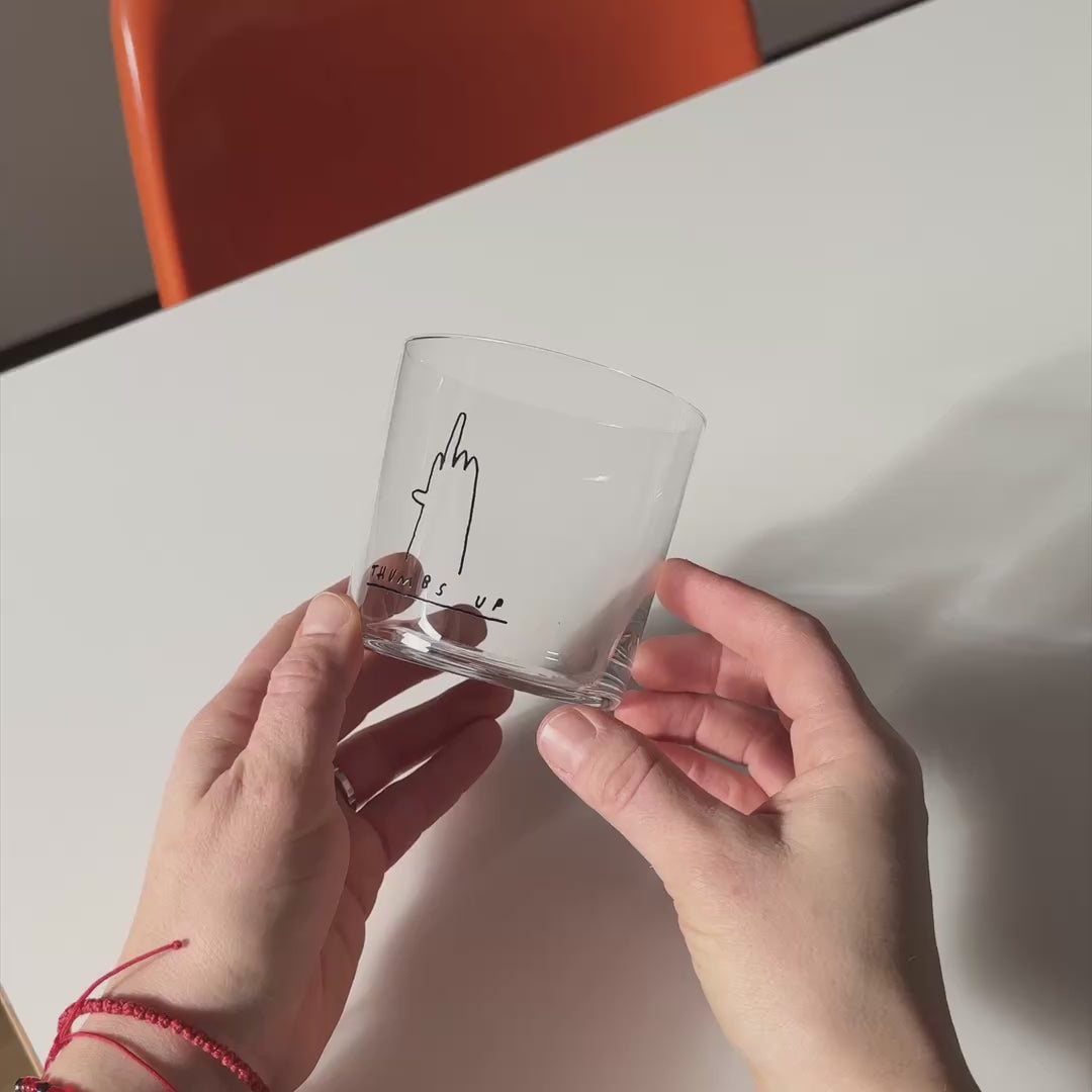 Medium Designer-Trinkglas mit sehr dünnen Glaswänden und einer schwarzen, einfarbigen Illustration auf der Vorderseite. Das Motiv zeigt eine Typografie in der geschrieben THUMBS UP und einer Illustration einer Hand die den Mittelfinger.

Das Glas wird über einem weißen Tisch  mit orangenen Stühlen von zwei Händen gehalten und langsam hin und her bewegt.