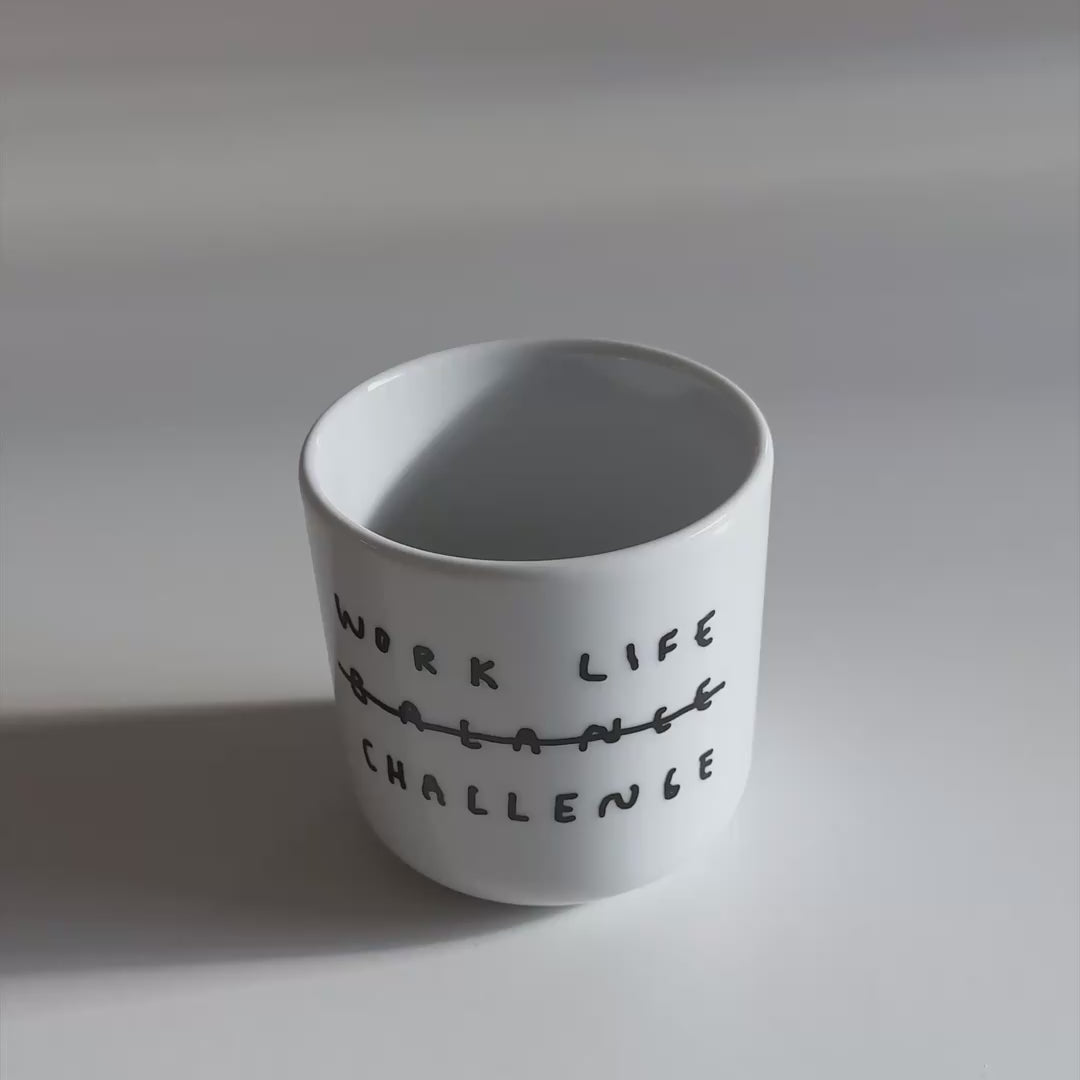 Weißer henkelloser Porzellan-Becher mit einer einfachen schwarzen Illustration, einem handschriftlichen Text der besagt "WORK LIFE CHALLENGE" wobei auch das Wort Balance dabeisteht und humorvoll durchgestrichen wird, dadurch von Challenge ersetzt wird. Der Becher steht auf weißen Untergrund und wird im VIdeo mit Cornflakes befüllt. 