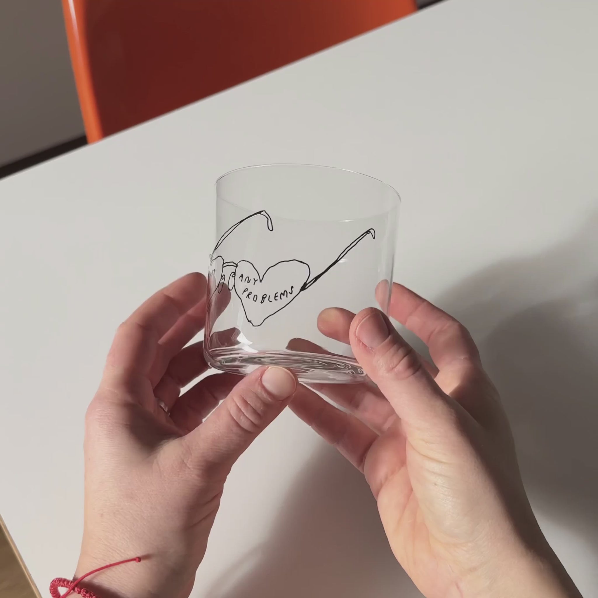 Transparentes Medium Designer Glas mit schwarzem Schriftzug "can't see any problems" mit einer Brille, die Brillengläser in herzformen hat. Das Glas wird in zwei Händen gehalten und hin und her bewegt, dahinter ein weißer Tisch und orangener Stuhl. 