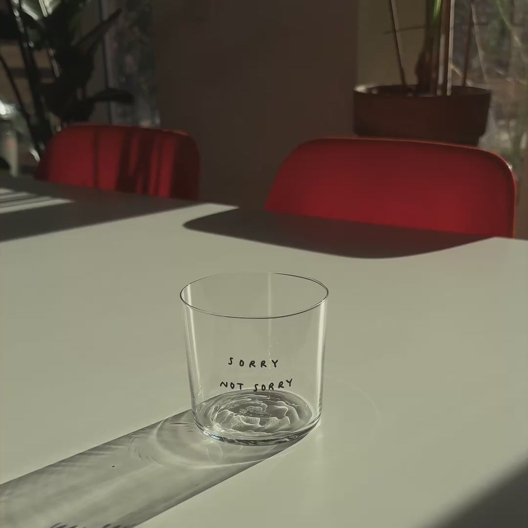 Medium Designer-Trinkglas mit sehr dünnen Glaswänden und einer schwarzen, einfarbigen Illustration auf der Vorderseite. Das Motiv zeigt eine Typografie in der geschrieben ist SORRY NOT SORRY.

Das Glas steht auf einem weißen Tisch in angenehmen Licht, dahinter stehen zwei orangene Stühle und mehrere Pflanzen. Das Glas wird mit Tee befüllt.