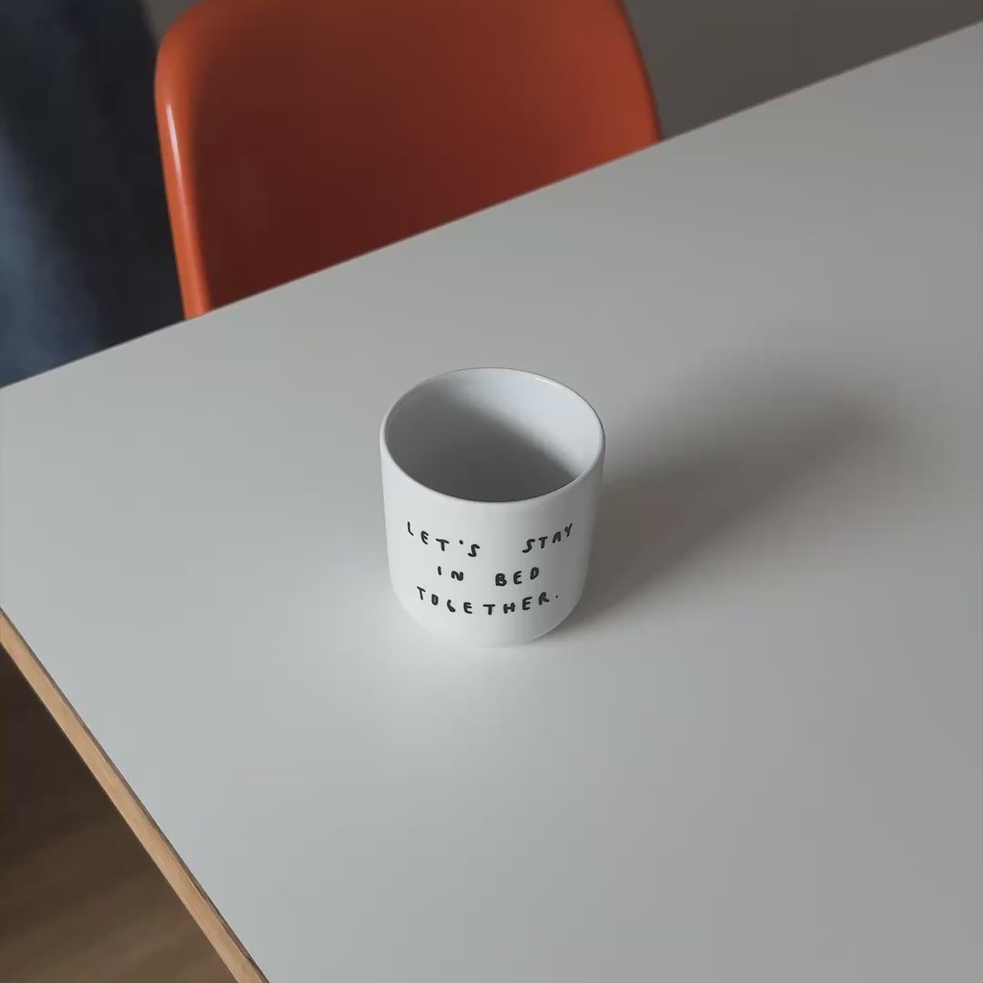 Weißer henkelloser Porzellan-Becher mit einer einfachen schwarzen Illustration, einem handschriftlichen Text der besagt "LET'S STAY IN BED TOGETHER" auf einem weißen Tisch mit orangenen Stuhl. Im Hintergrund läuft eine Person die im Video Tee in den Becher füllt.