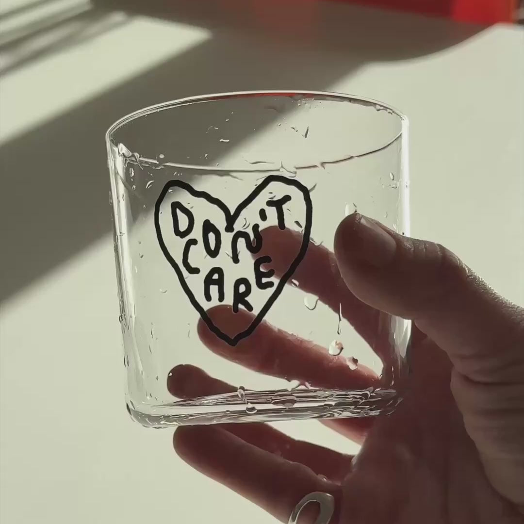 Transparentes Medium Designer Glas mit schwarzem Schriftzug "DON'T CARE" in einem Herz mit dünnen Wänden und Boden. 

Das Glas ist nass und wird von einer Hand gehalten vor einem weißen Tisch. Das Glas wird in Nahaufnahme hin und her bewegt und im VIdeo abgetrocknet.