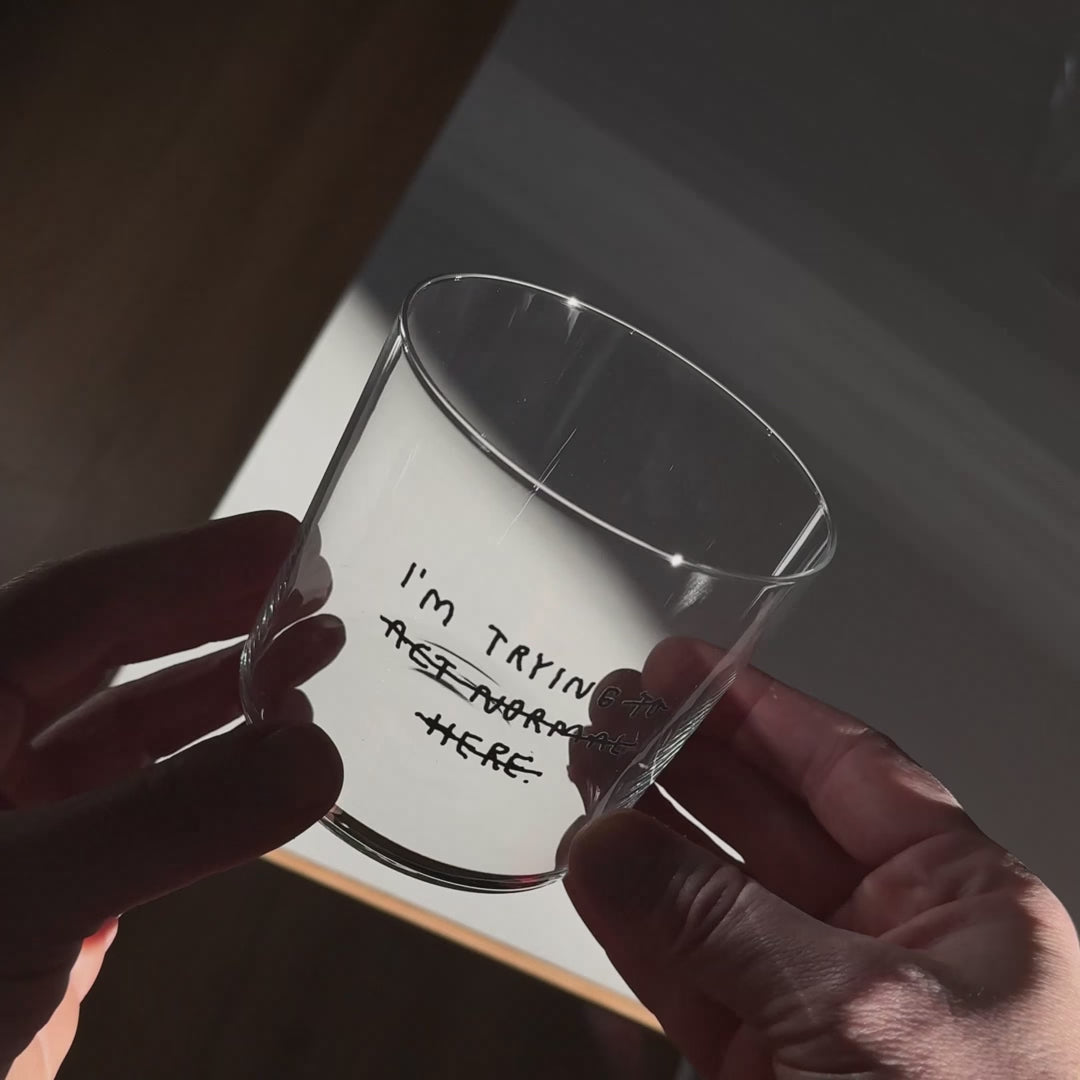 Transparentes Medium Designer Glas mit schwarzem Schriftzug "i'm trying to act normal here" mit dünnen Wänden und Boden. Dabei ist das "to act normal here durchgestrichen, so dass lediglich die Aussage "I'm trying" steht.

Das Glas ist mit einer transparenten Flüssigkeit befüllt und Eiswürfeln. Es steht auf einem weißen Tisch und wirft einen schatten.