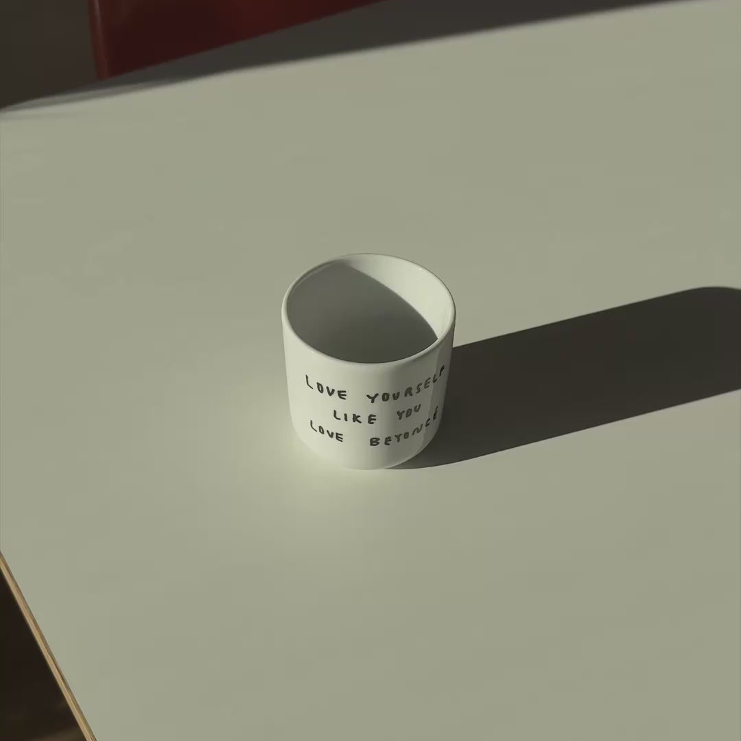 Weißer henkelloser Porzellan-Becher  mit einer einfachen schwarzen Illustration, einem handschriftlichen Text der besagt "LOVE YOURSELF LIKE YOU LOVE BEYONCÉ" auf einem weißen Tisch. Der Becher wird in dem Video von zwei Händen hochgehoben und hin und her bewegt. 