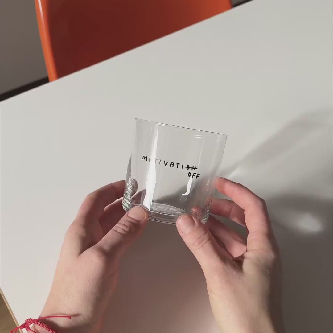 Transparentes Designer Glass mit filigranen dünnen Wänden und Boden schwarzem Schriftzug "Motivatioff", wobei eigentlich Motivation geschrieben ist, das ON am Ende durchgestrichen ist, und durch OFF ersetzt wird. Weißer Hintergrund.

Das Glas wird von zwei Händen über einem weißen Tisch gehalten und langsam hin und her bewegt. 