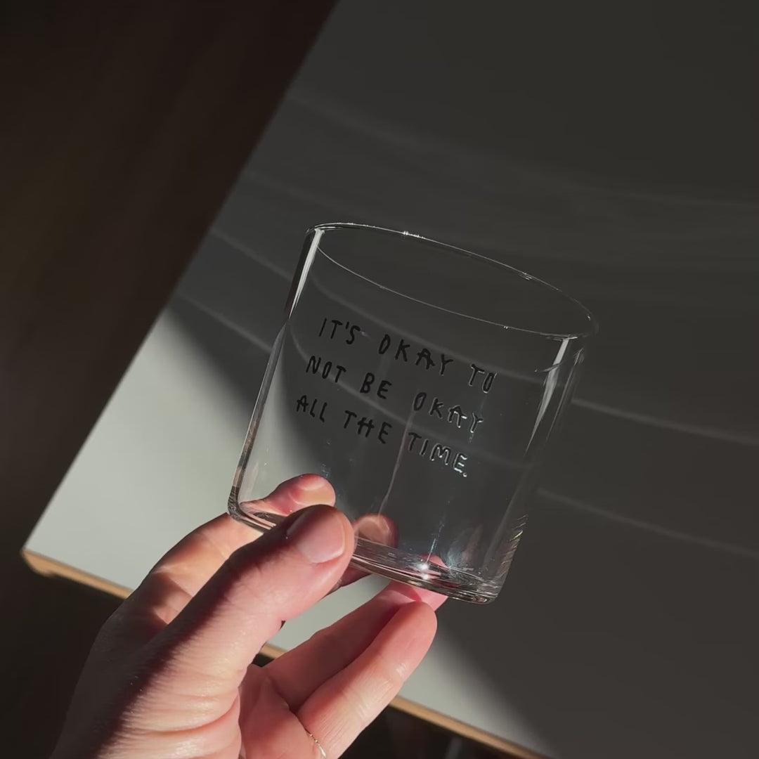 Transparentes Medium Designer Glas mit schwarzem Schriftzug "It's okay to not be okay all the time" mit dünnen Wänden und Boden. 

Das Glas ist mit einer transparenten Flüssigkeit befüllt. Es steht auf einem weißen Tisch und wirft einen schatten. Daneben steht ein orangener Stuhl.