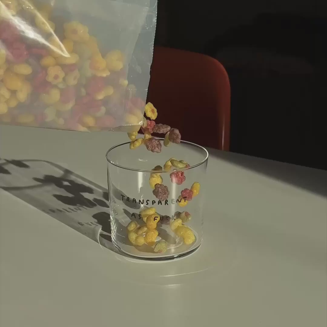 Medium Designer-Trinkglas mit sehr dünnen Glaswänden und einer schwarzen, einfarbigen Illustration auf der Vorderseite. Das Motiv zeigt eine Typografie in der geschrieben ist TRANSPARENT AS FUCK.

Das Glas steht auf einem weißen Tisch mit orangenen Stuhl im Hintergrund. Das Glas wird mit bunten Cornflakes befüllt. 