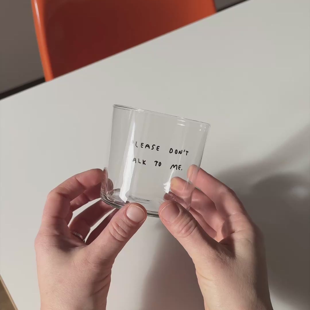 Medium Designer-Trinkglas mit sehr dünnen Glaswänden und einer schwarzen, einfarbigen Illustration auf der Vorderseite. Das Motiv zeigt eine Typografie in der geschrieben ist PLEASE DON'T TALK TO ME.

Das Glas wird über einem Weißen Tisch mit orangenen Stuhl in zwei Händen gehalten und langsam hin und her bewegt. 