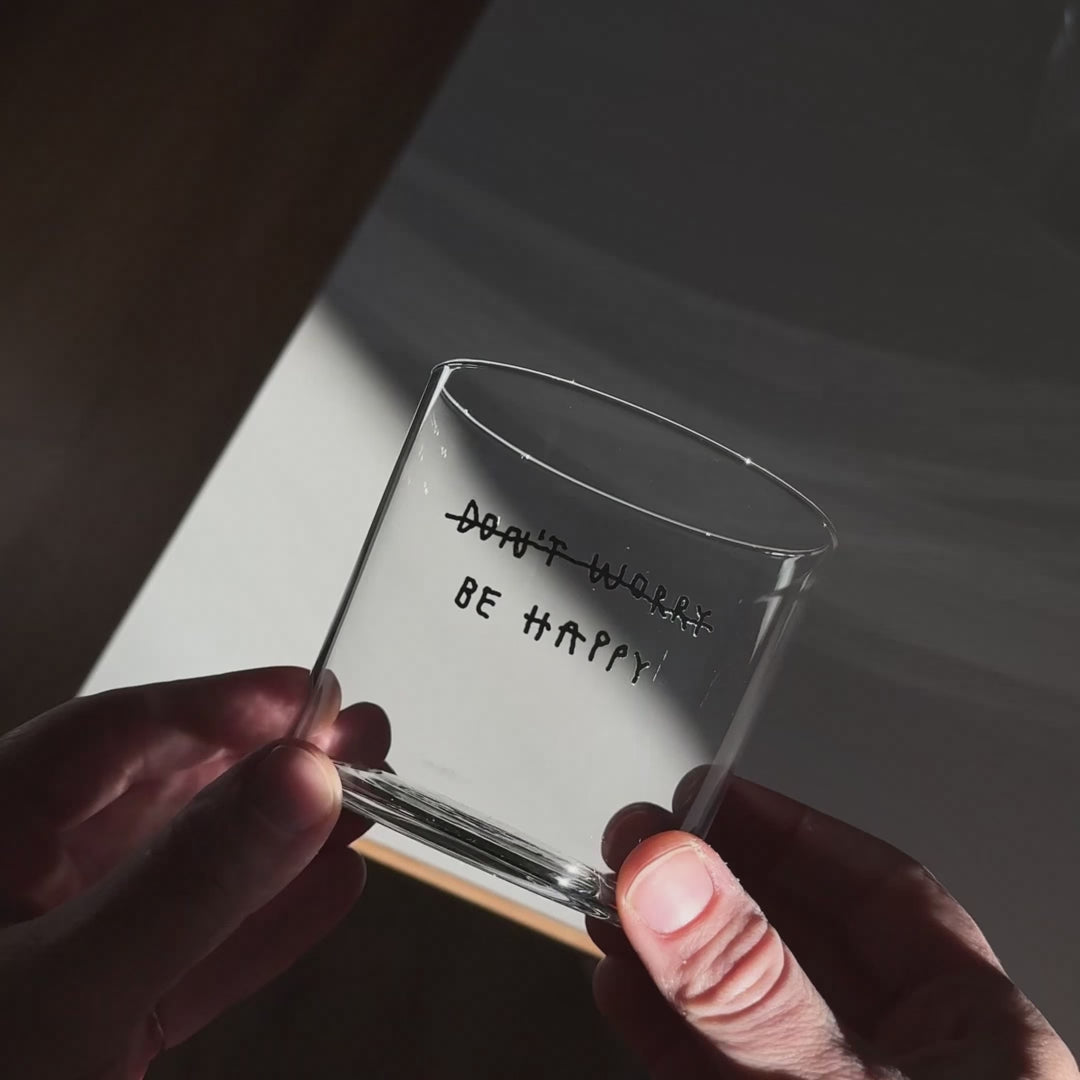 Transparentes Medium Designer Glas mit schwarzem Schriftzug "DONT WORRY BE HAPPY" wobei DONT WORRY durchgestrichen ist und lediglich die Aufforderung BE HAPPY im Vordergrund steht, die Gläser haben dünnen Wänden und Boden. 
 
Das Glas ist mit unbefüllt, das Foto wird von oben geschossen. Das Motiv ist lediglich als Schattenwurf im Glas sichtbar. 