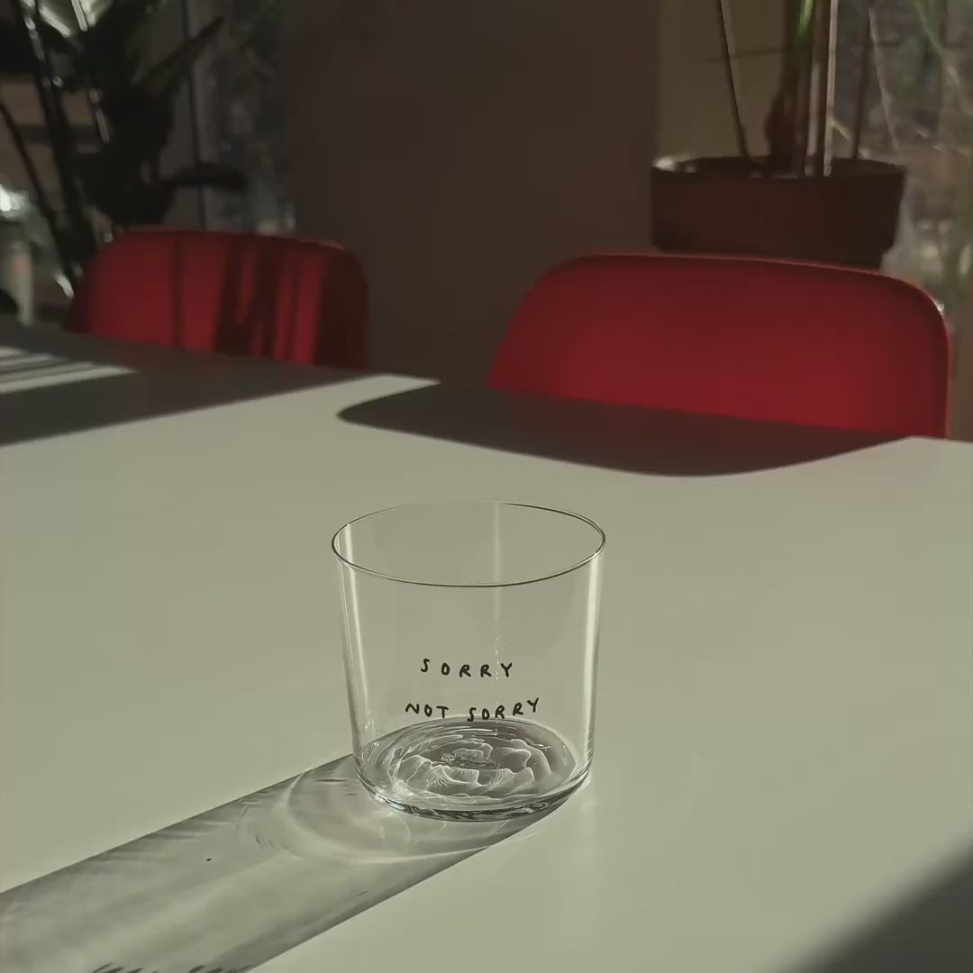 Medium Designer-Trinkglas mit sehr dünnen Glaswänden und einer schwarzen, einfarbigen Illustration auf der Vorderseite. Das Motiv zeigt eine Typografie in der geschrieben ist SORRY NOT SORRY.

Das Glas steht auf einem weißen Tisch in angenehmen Licht, dahinter stehen zwei orangene Stühle und mehrere Pflanzen. Das Glas wird mit Tee befüllt.