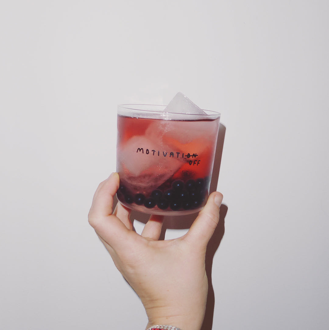 Transparentes Designer Glass mit filigranen dünnen Wänden und Boden schwarzem Schriftzug "Motivatioff", wobei eigentlich Motivation geschrieben ist, das ON am Ende durchgestrichen ist, und durch OFF ersetzt wird. Weißer Hintergrund.

Das Glas ist mit einer roten Flüssigkeit, Eiswürfeln und Blaubeeren befüllt. Das Glas wird von einer Hand hochgehalten vor einer weißen Wand.