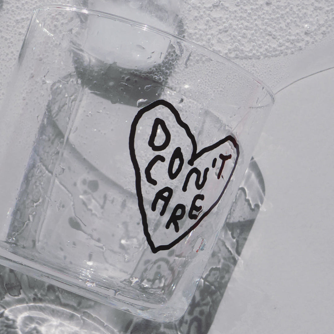 Transparentes Medium Designer Glas mit schwarzem Schriftzug "DON'T CARE" in einem Herz mit dünnen Wänden und Boden. 

Nahaufnahme des feuchten Glasses welches auf einem weißen Tisch liegt. Aus dem Glas läuft Wasser und ein Eiswürfel.